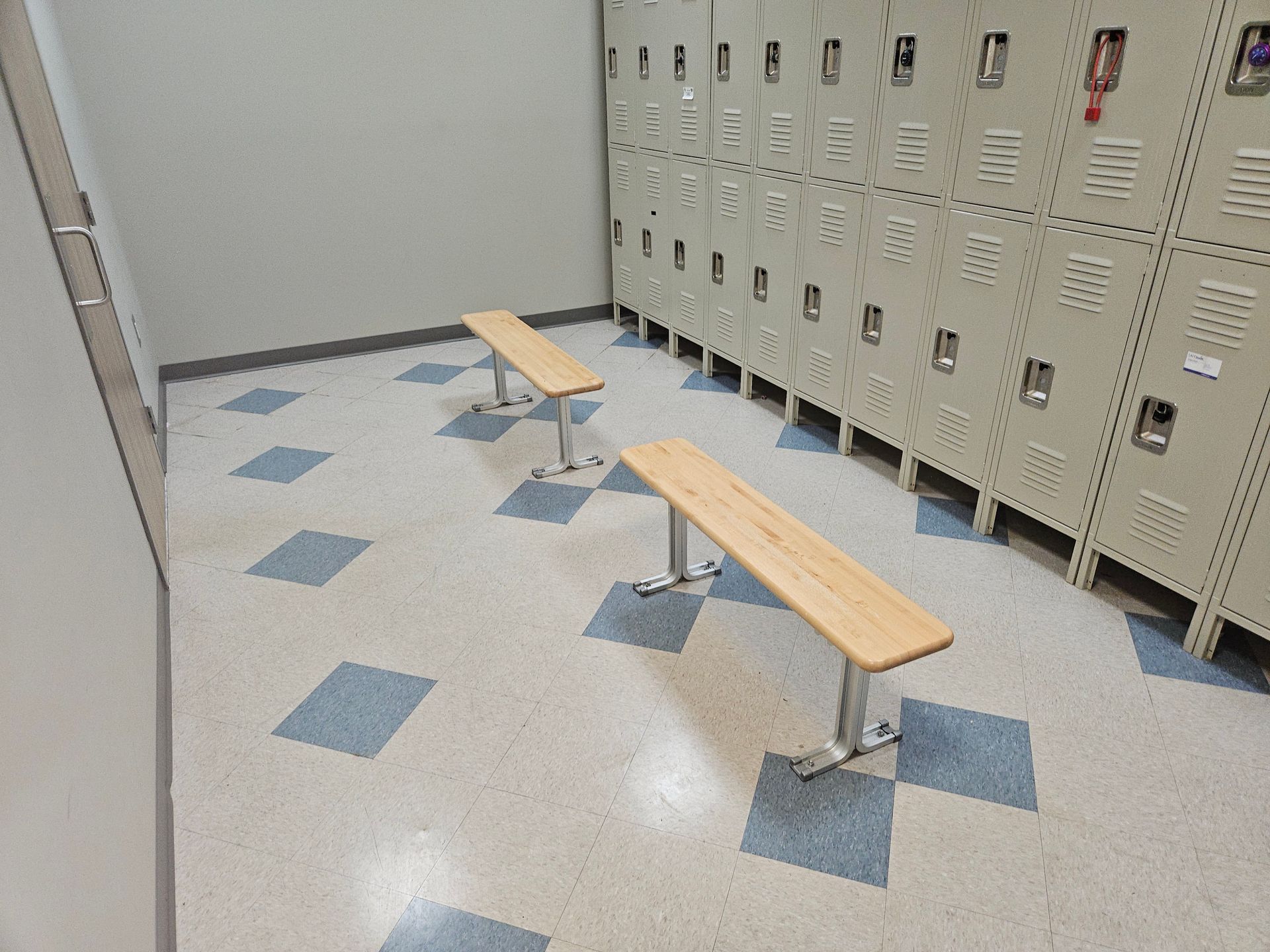 Locker room with two wooden benches on a blue-and-gray tiled floor beside rows of beige lockers.