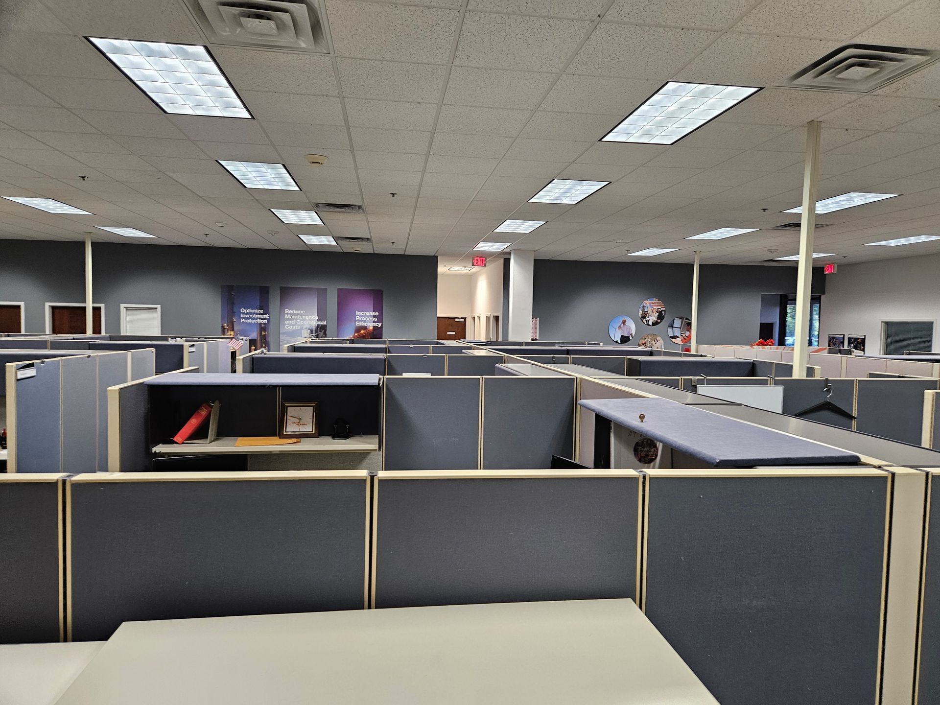 Empty office cubicles under fluorescent ceiling lights in a gray, open-plan workspace.
