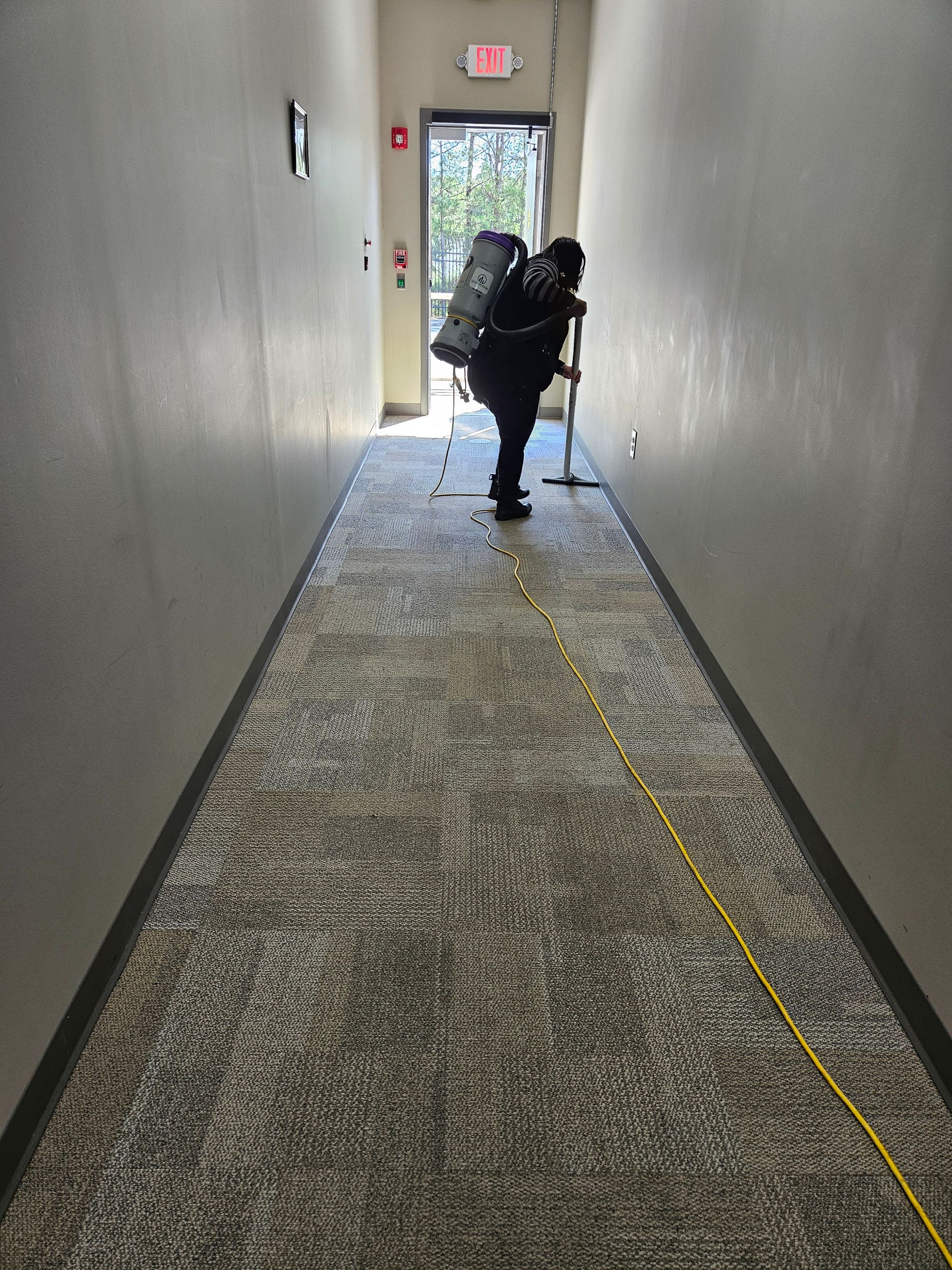 Person with backpack walking down a narrow carpeted hallway toward an exit door.