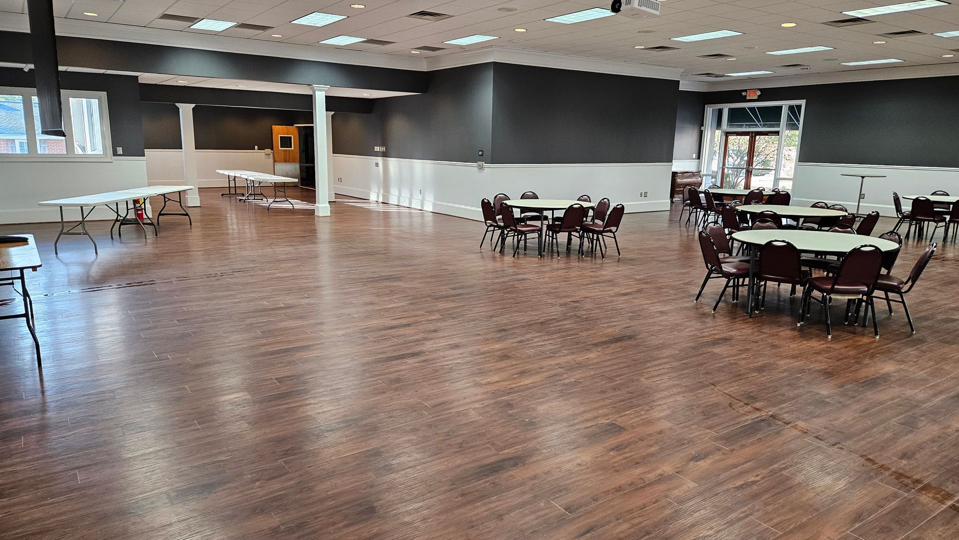 Empty event hall with polished wooden floor, round tables, and chairs under bright indoor lighting