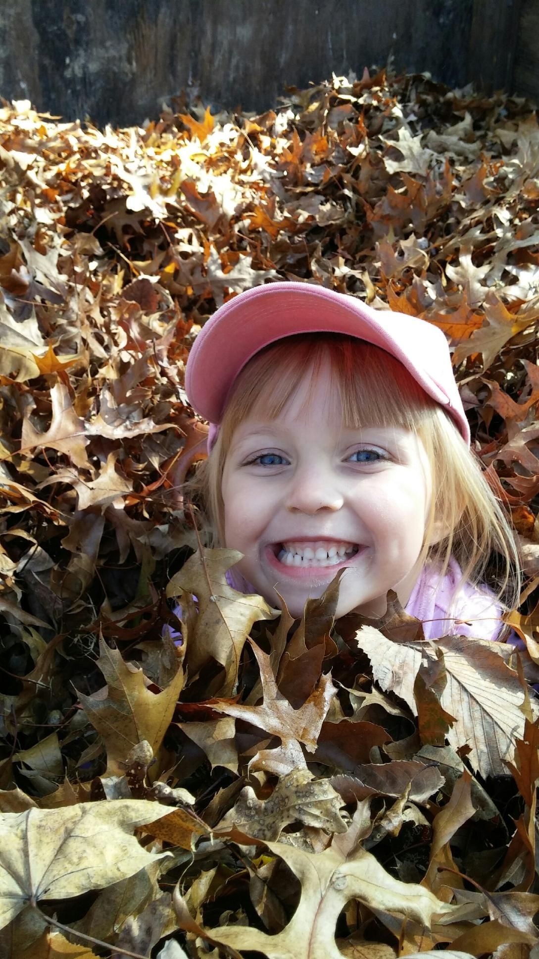 Child smiling in a pile of autumn leaves, wearing a pink cap.