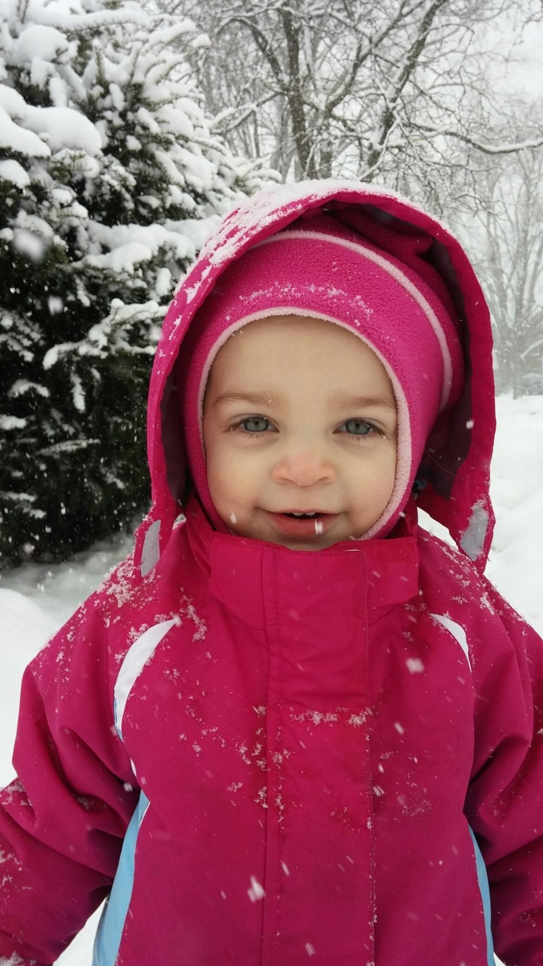 Child in a bright pink snowsuit standing in falling snow near snow-covered trees