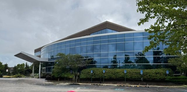 Modern glass office building with curved facade and peaked roof, viewed from the parking lot.