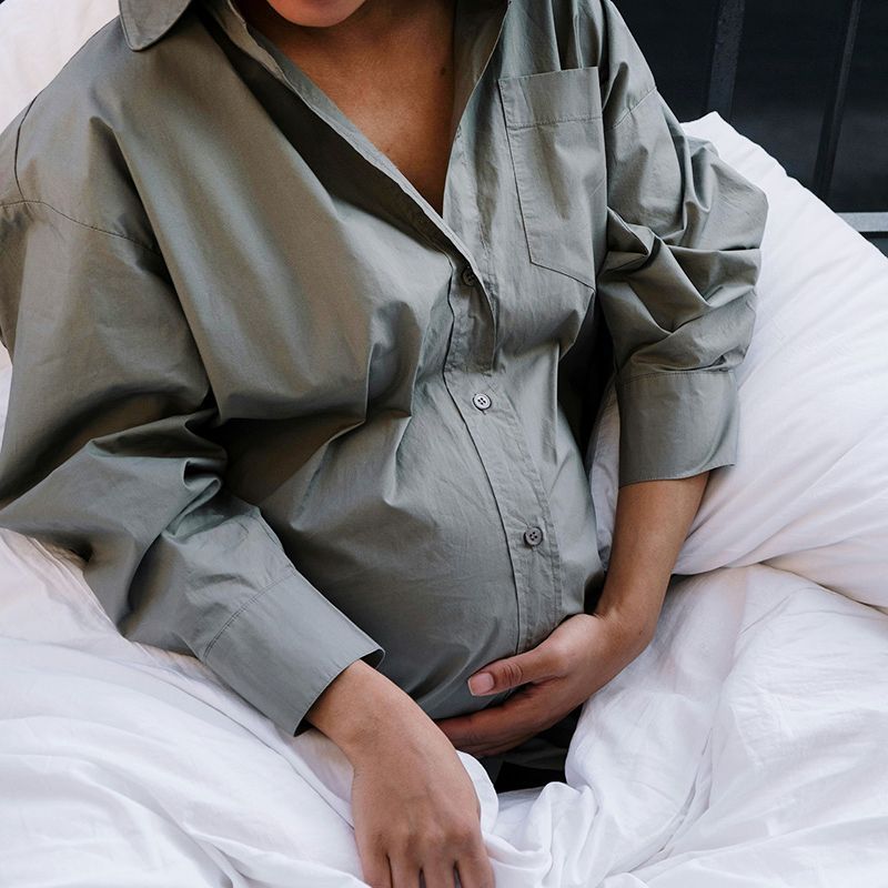 Pregnant person in a gray shirt sitting on a bed, hands resting on belly