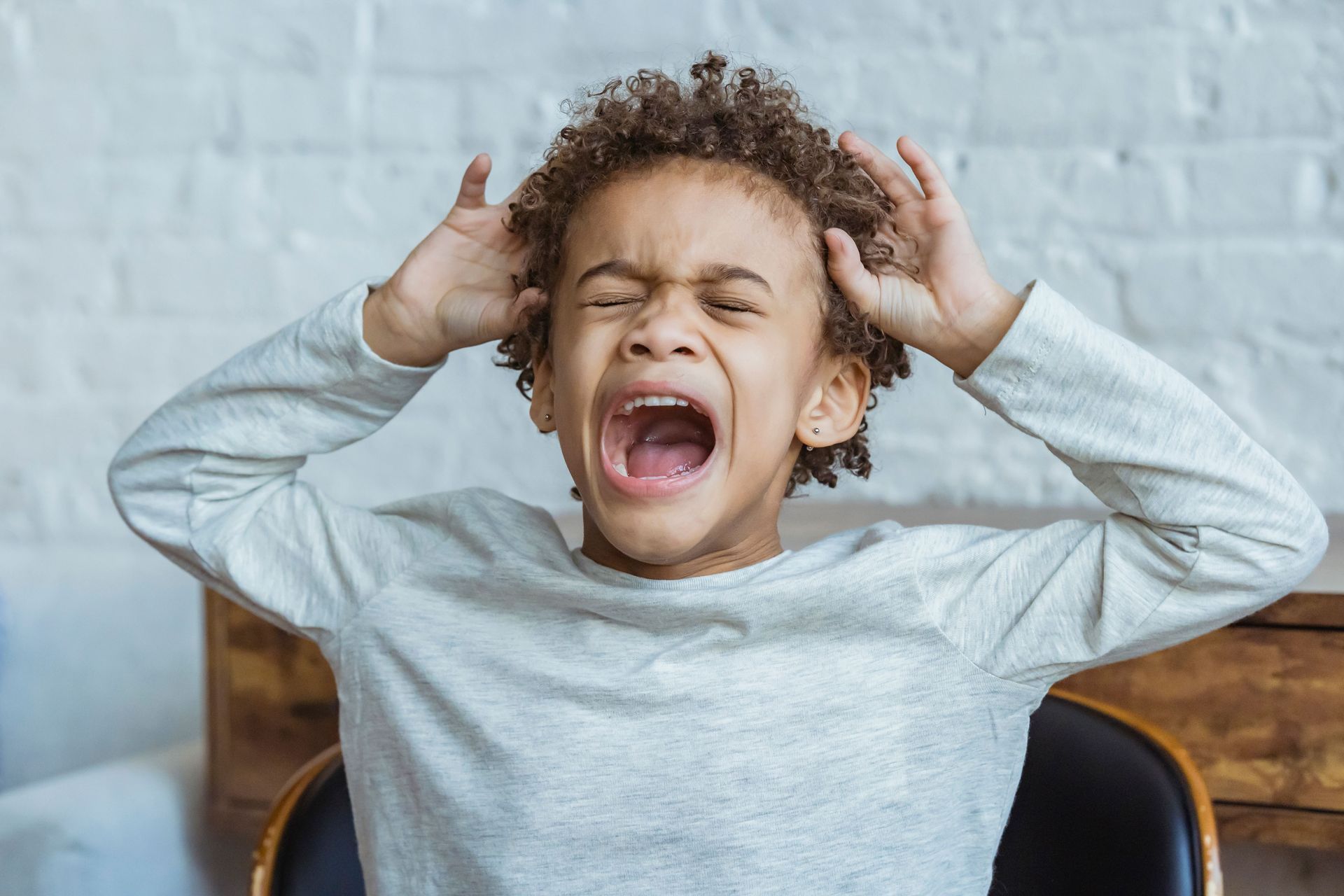 Child with curly hair yawning loudly, hands near ears, wearing a gray shirt on a bed