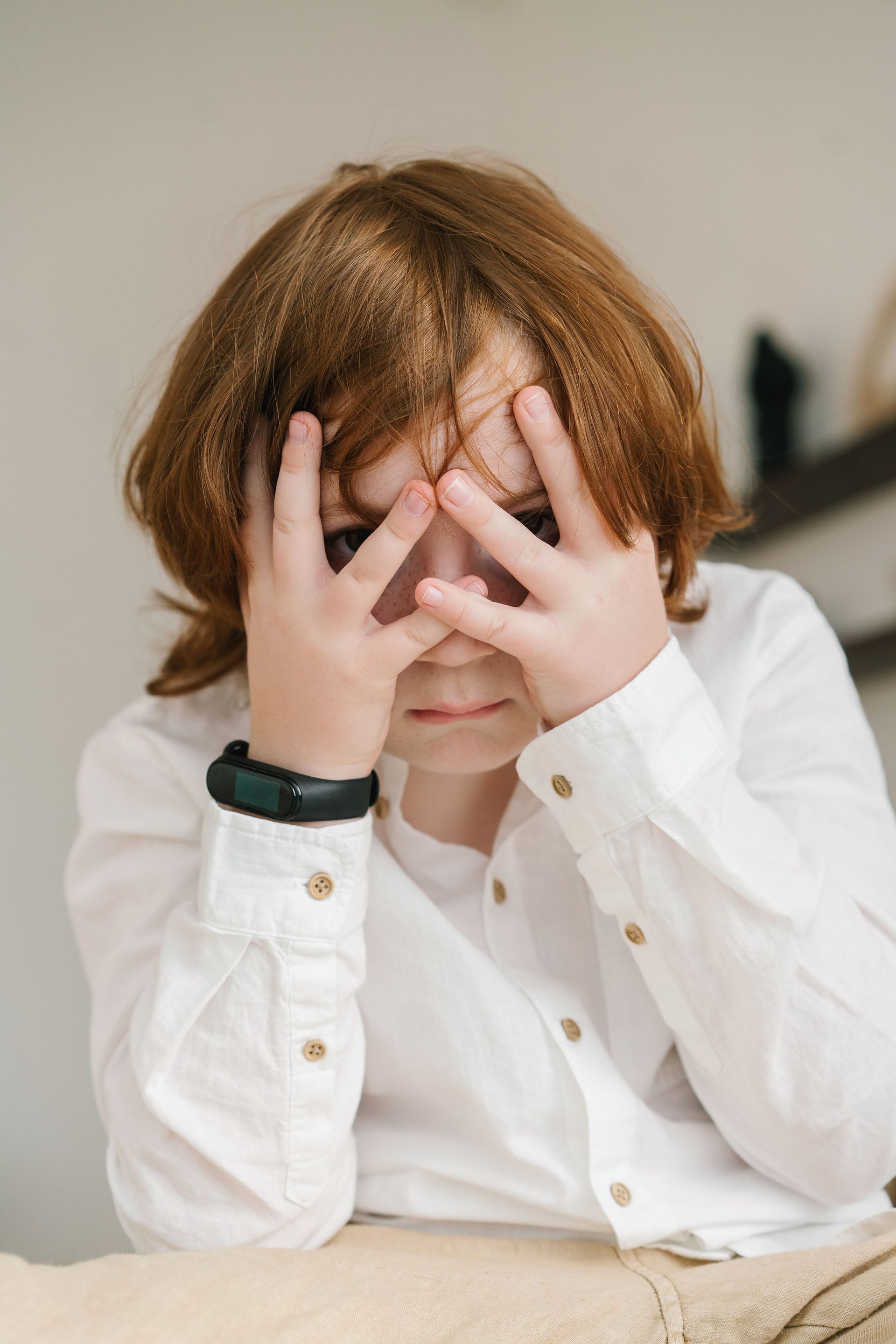Child covering face with both hands while sitting at a table, wearing a white shirt and black wristband.