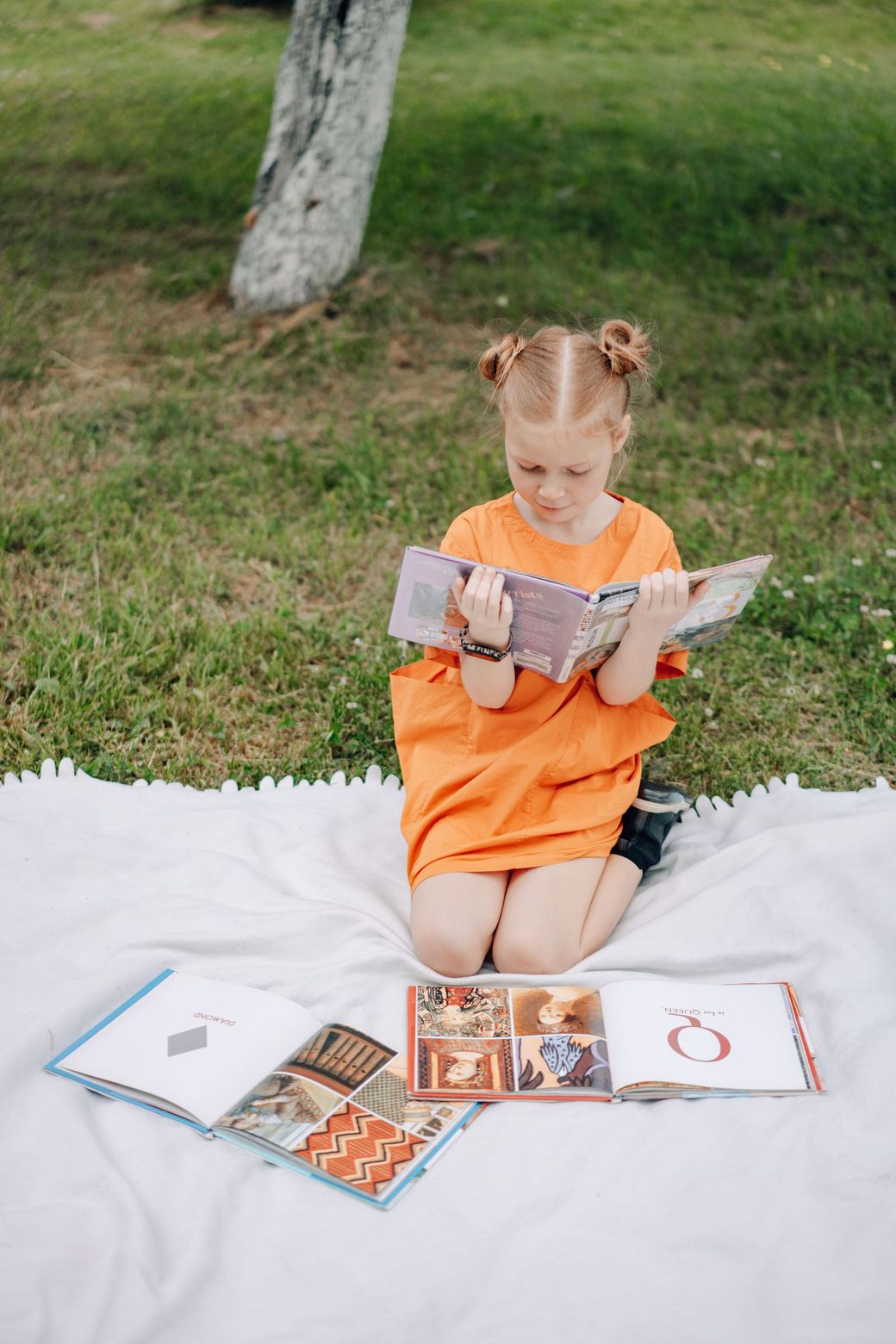 Child in an orange outfit reading a book on a white blanket outdoors, with several books spread around.