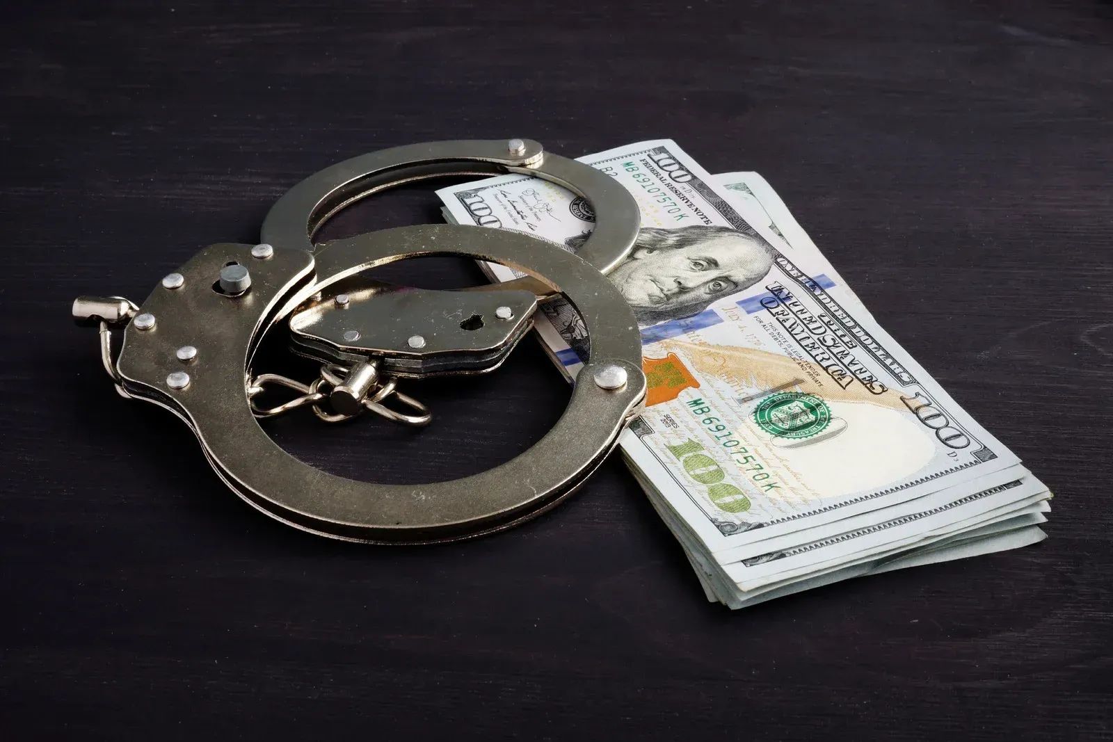 Handcuffs beside a stack of U.S. dollar bills on a dark surface
