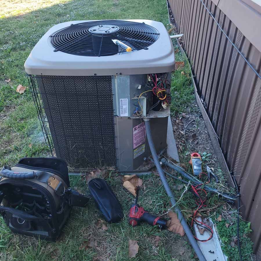 Damaged outdoor air conditioner with open panel and tools on grass beside a fence