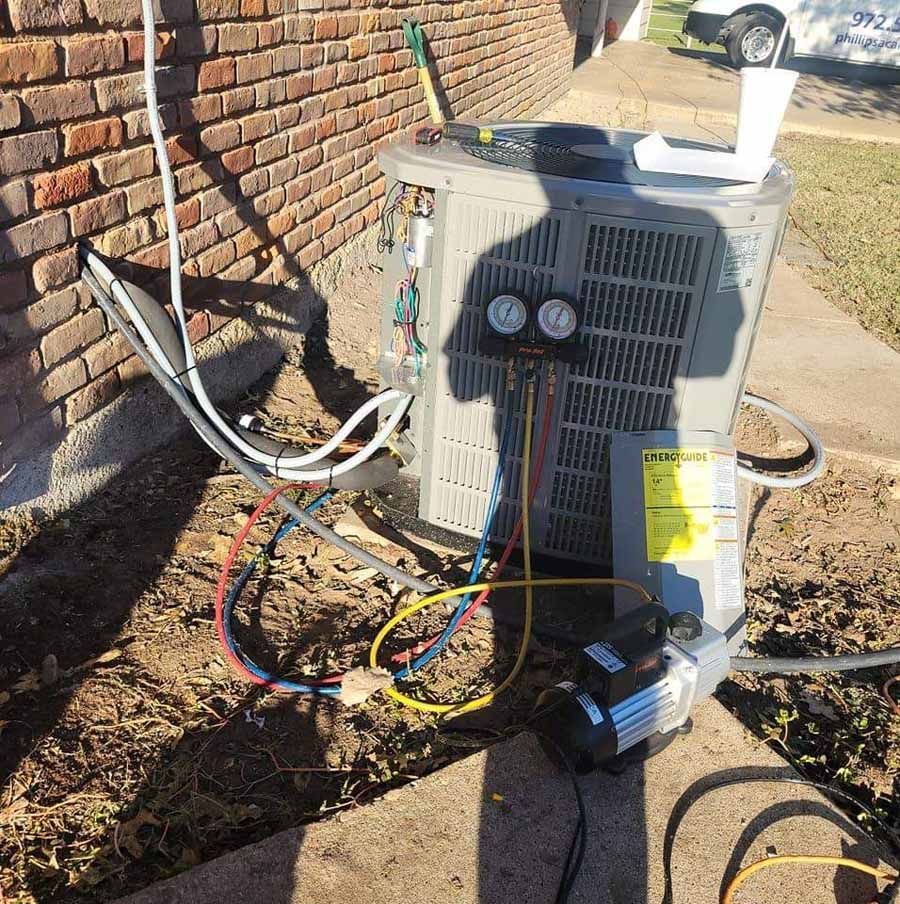 Outdoor HVAC unit with exposed wiring and gauges beside a brick wall, with a detached component on the ground