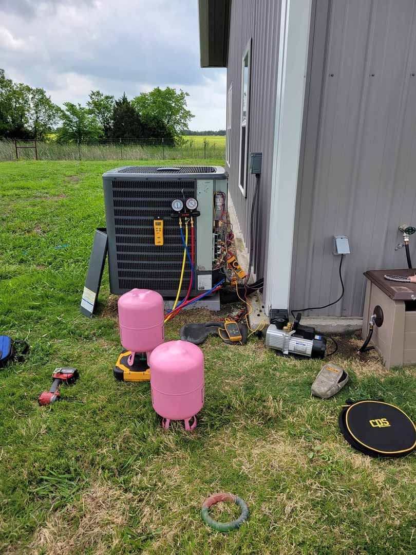 Outdoor HVAC unit beside a gray building, with pink tanks and tools on grass nearby.