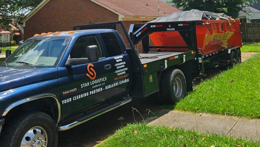 A blue flatbed truck with orange branding parked on a lawn, hauling an orange dumpster trailer covered with a tarp.