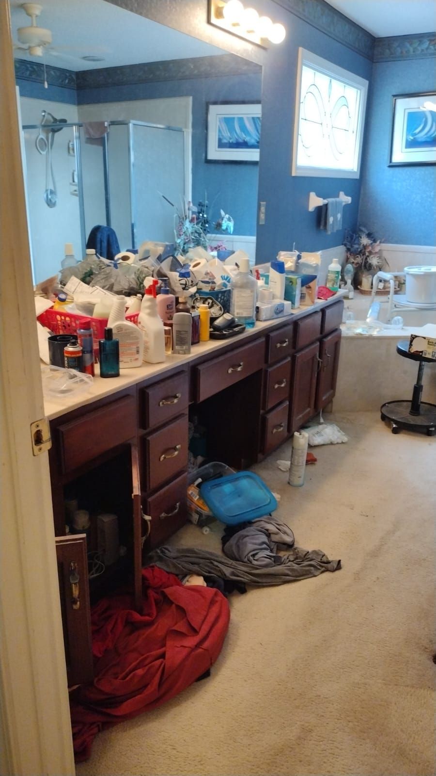 A cluttered bathroom counter with various toiletries, open cabinets, and items strewn on the carpeted floor.
