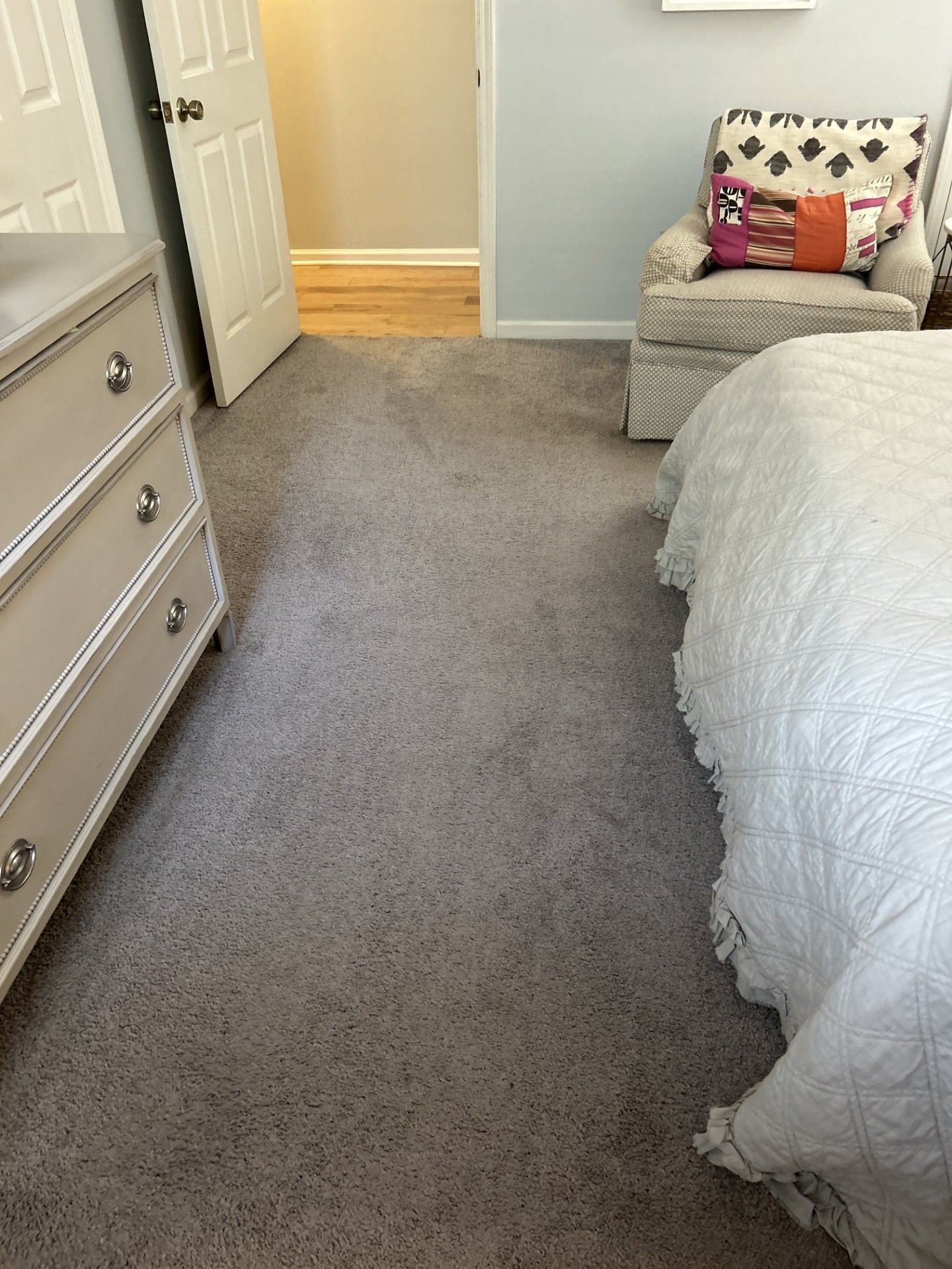 A bedroom with gray carpet, a white dresser on the left, a partially visible white bed, and an armchair in the corner.