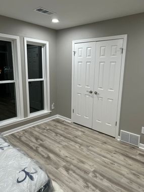 A room with light wood-look flooring, gray walls, a window on the left, and a closed white double door on the right.