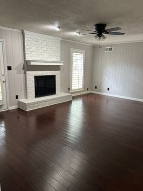 A living room with dark hardwood floors, grey patterned wallpaper, and a white brick fireplace with a wood mantel.