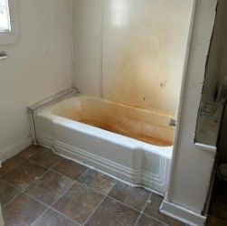 A stained, cream-colored bathtub with white surrounding wall panels sits on a tiled floor in a bathroom.