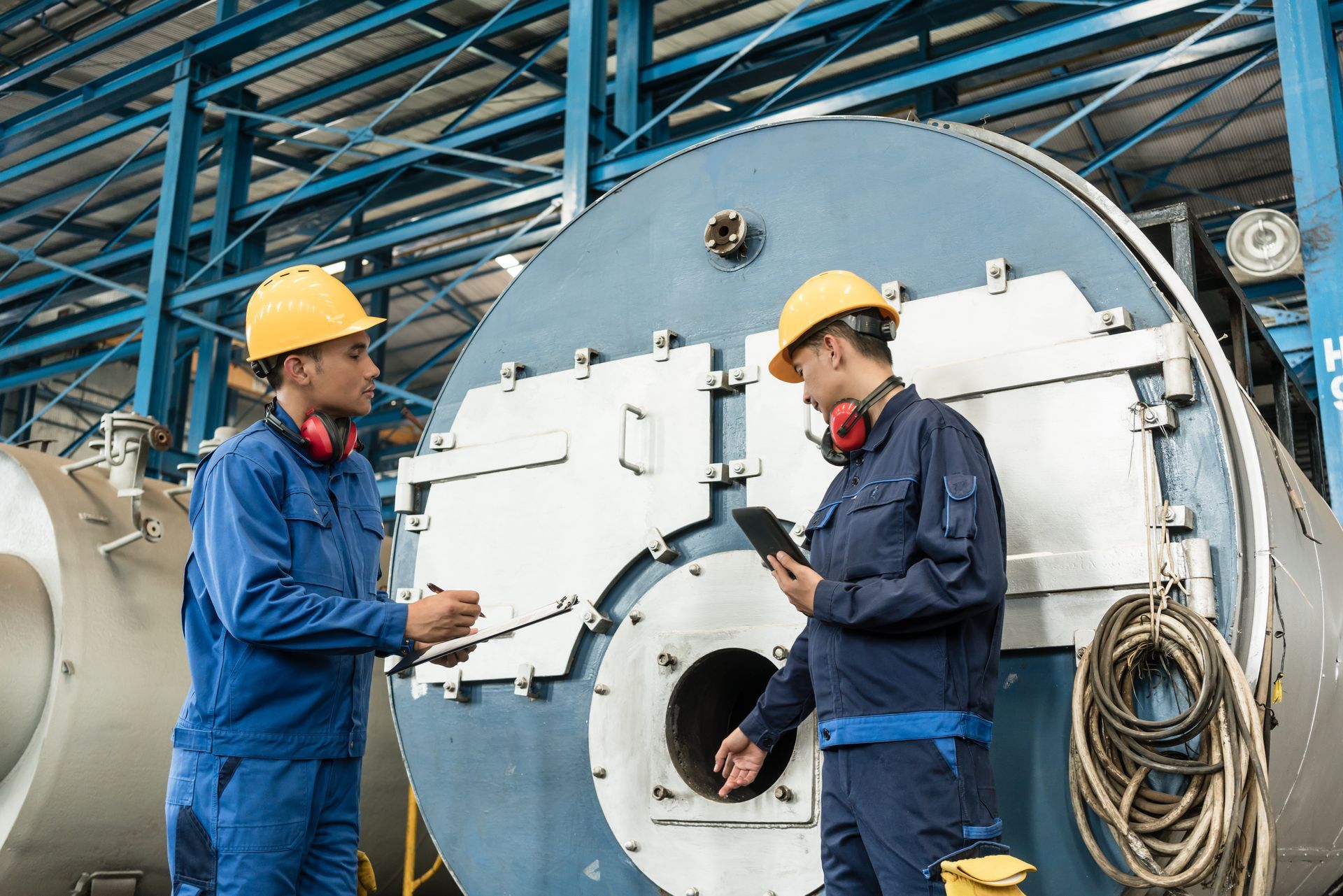 Two workers in hard hats inspecting a large industrial boiler in a plant.