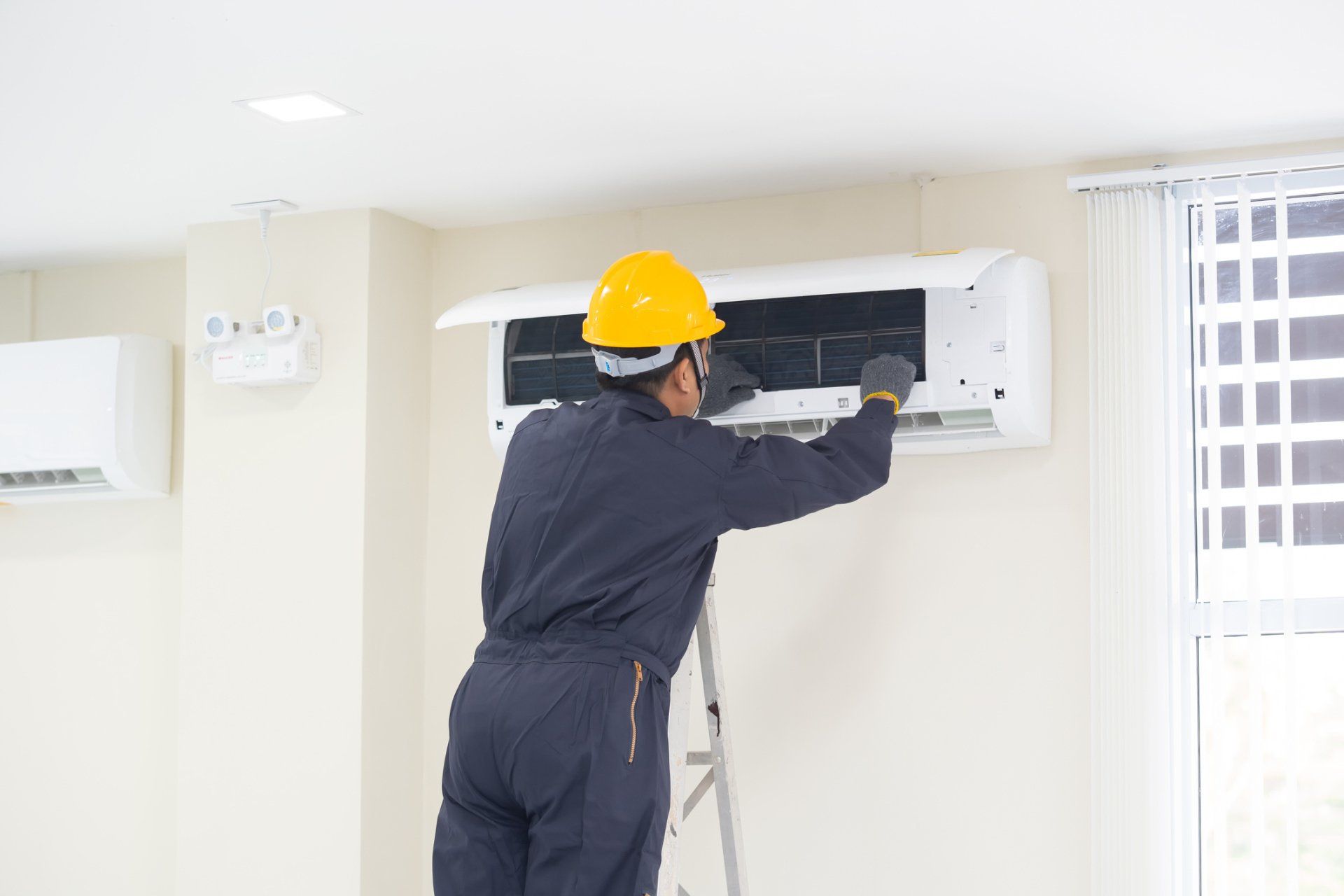Technician in yellow hard hat servicing a wall-mounted air conditioner indoors.