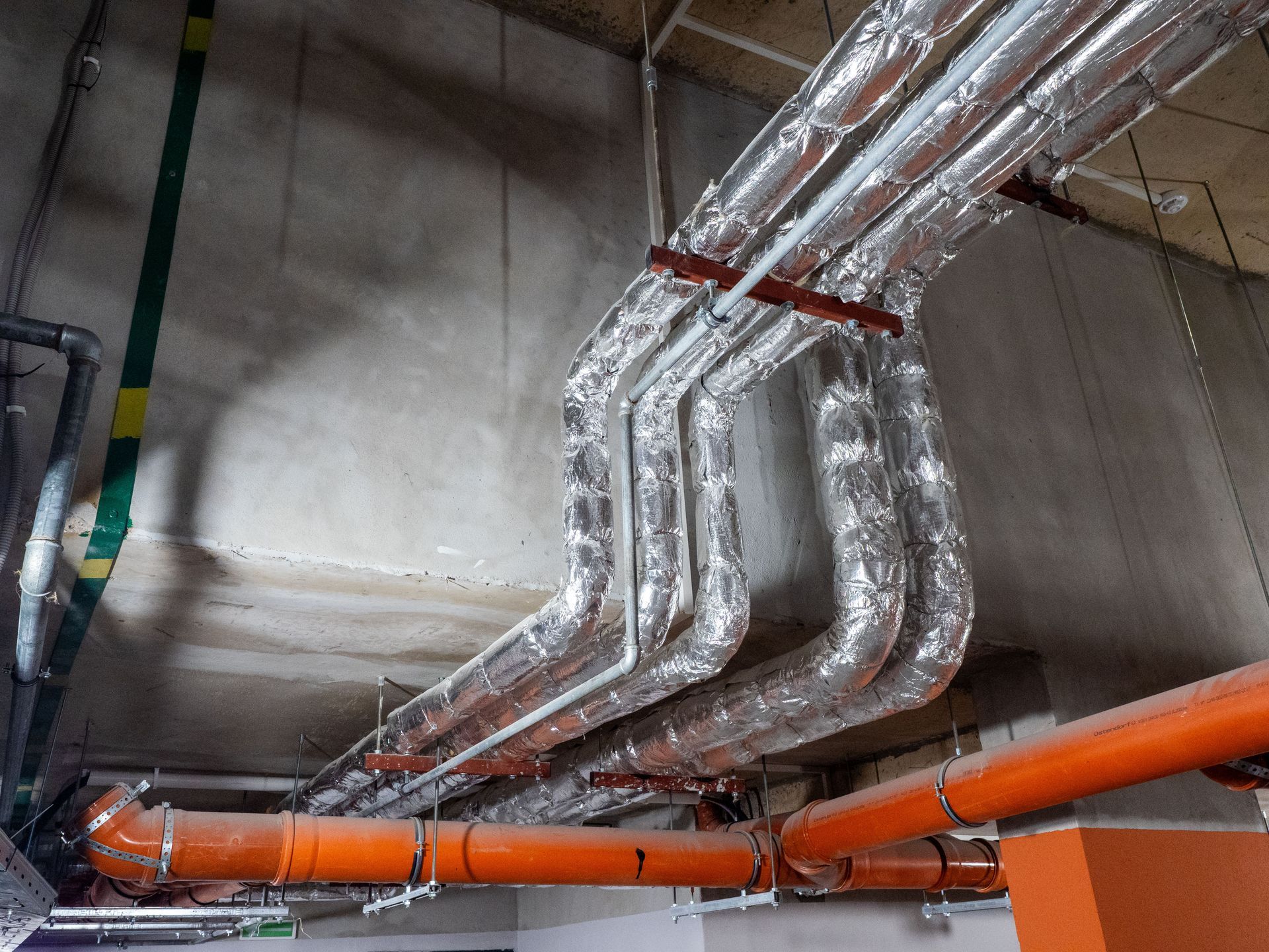 Insulated silver pipes and orange ducting run across a concrete industrial ceiling Insulated silver pipes and orange ducting run across a concrete industrial ceiling