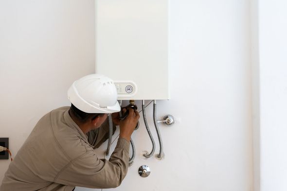 Worker installing plumbing under a wall-mounted cabinet, with exposed pipes and a white hard hat