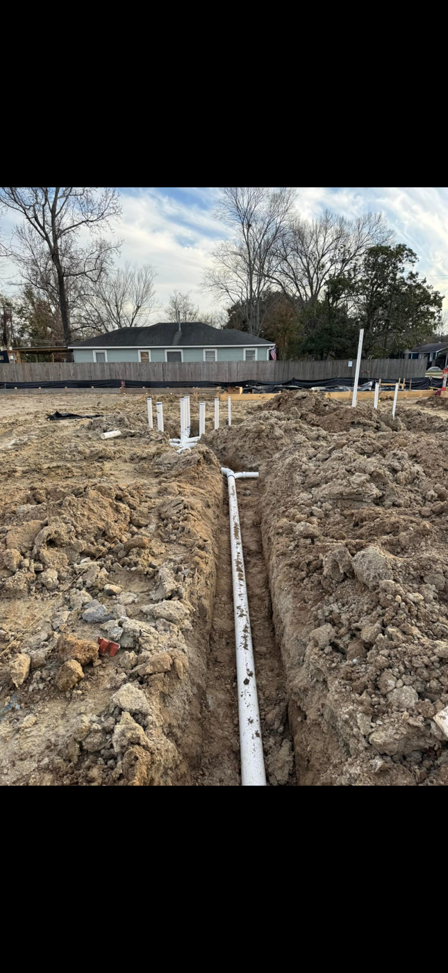 Construction trench with a white pipe running toward a house under renovation in a dirt yard