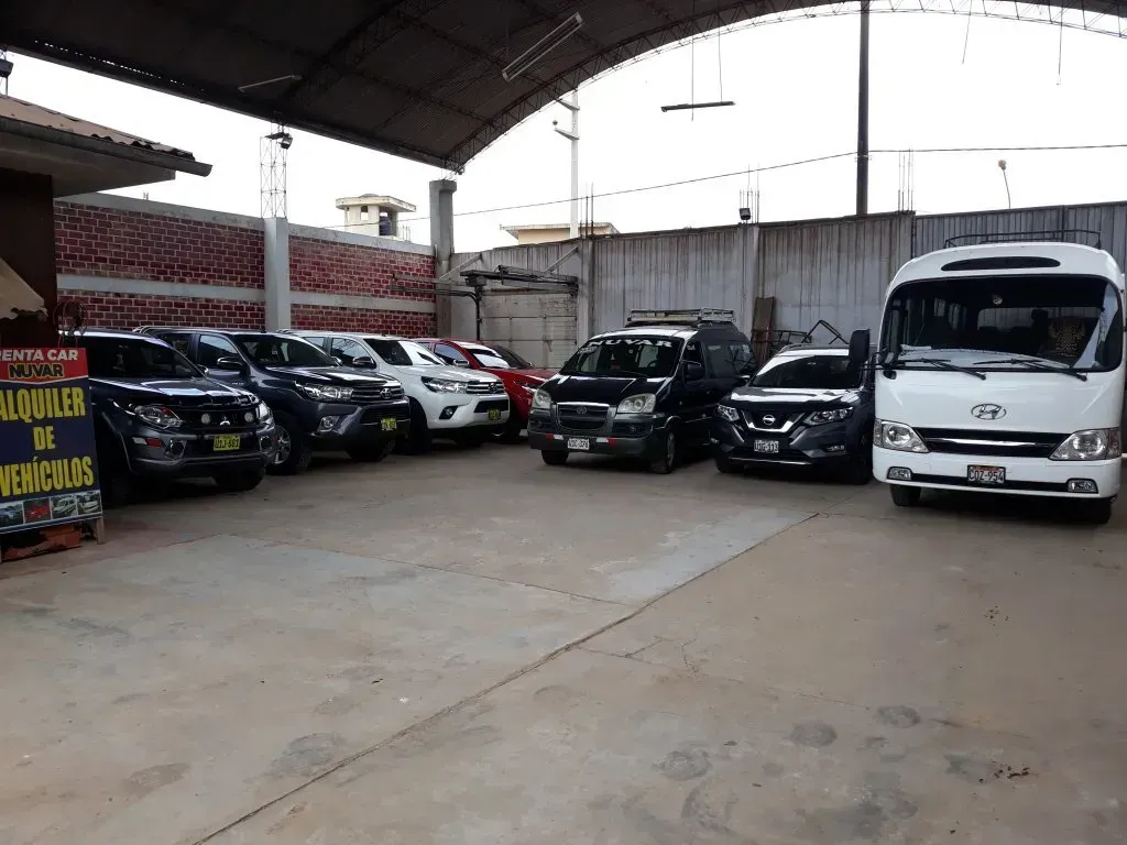 Aparcamiento cubierto con filas de coches aparcados y una furgoneta blanca bajo un gran techo.