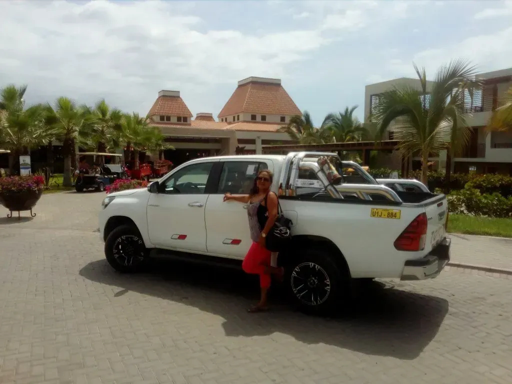Camioneta blanca con ruedas negras estacionada afuera de un complejo turístico, con una mujer posando junto a la puerta.