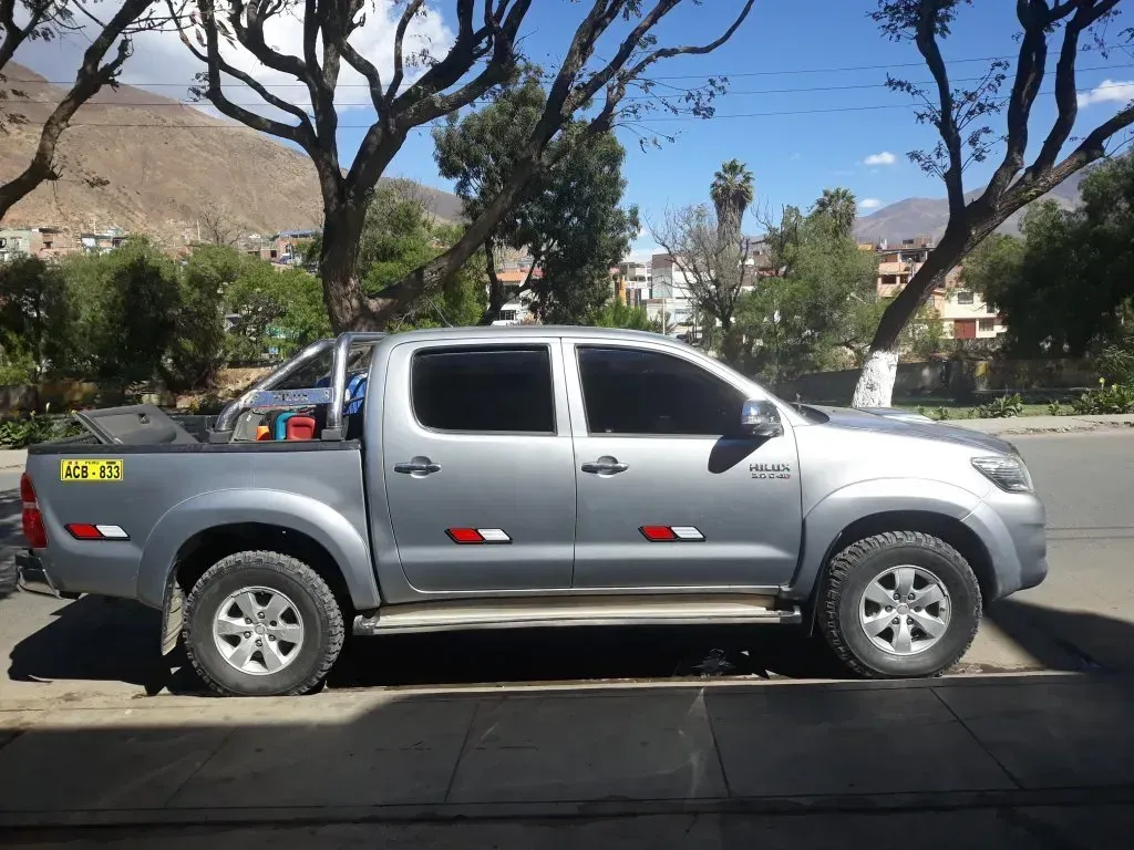 Camioneta plateada estacionada en una calle con árboles y colinas al fondo.