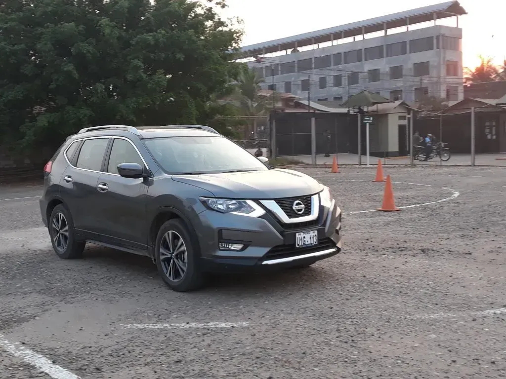 Un SUV gris estacionado en un terreno de grava cerca de conos naranjas, con un edificio y árboles al fondo.
