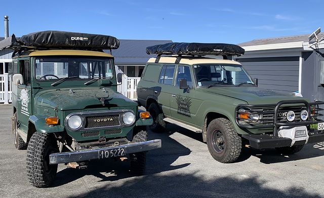 Two green off-road vehicles parked side by side, two classic Land Cruisers FJ40 & FJ60 with rooftop gear.