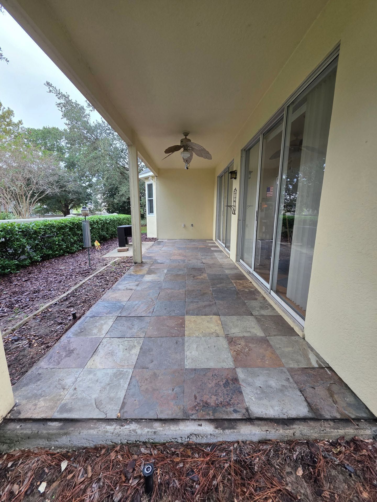 Covered patio with stone tile floor, ceiling fans, and sliding glass doors beside landscaped greenery