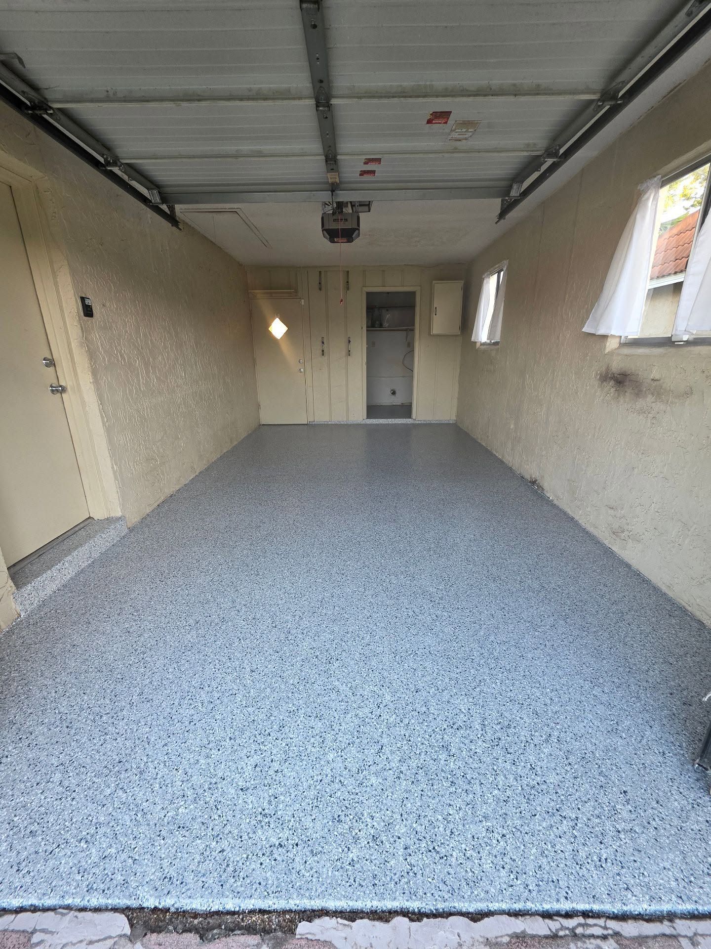 Empty garage with speckled blue-gray floor, beige walls, closed door at the back, and windows along the right side