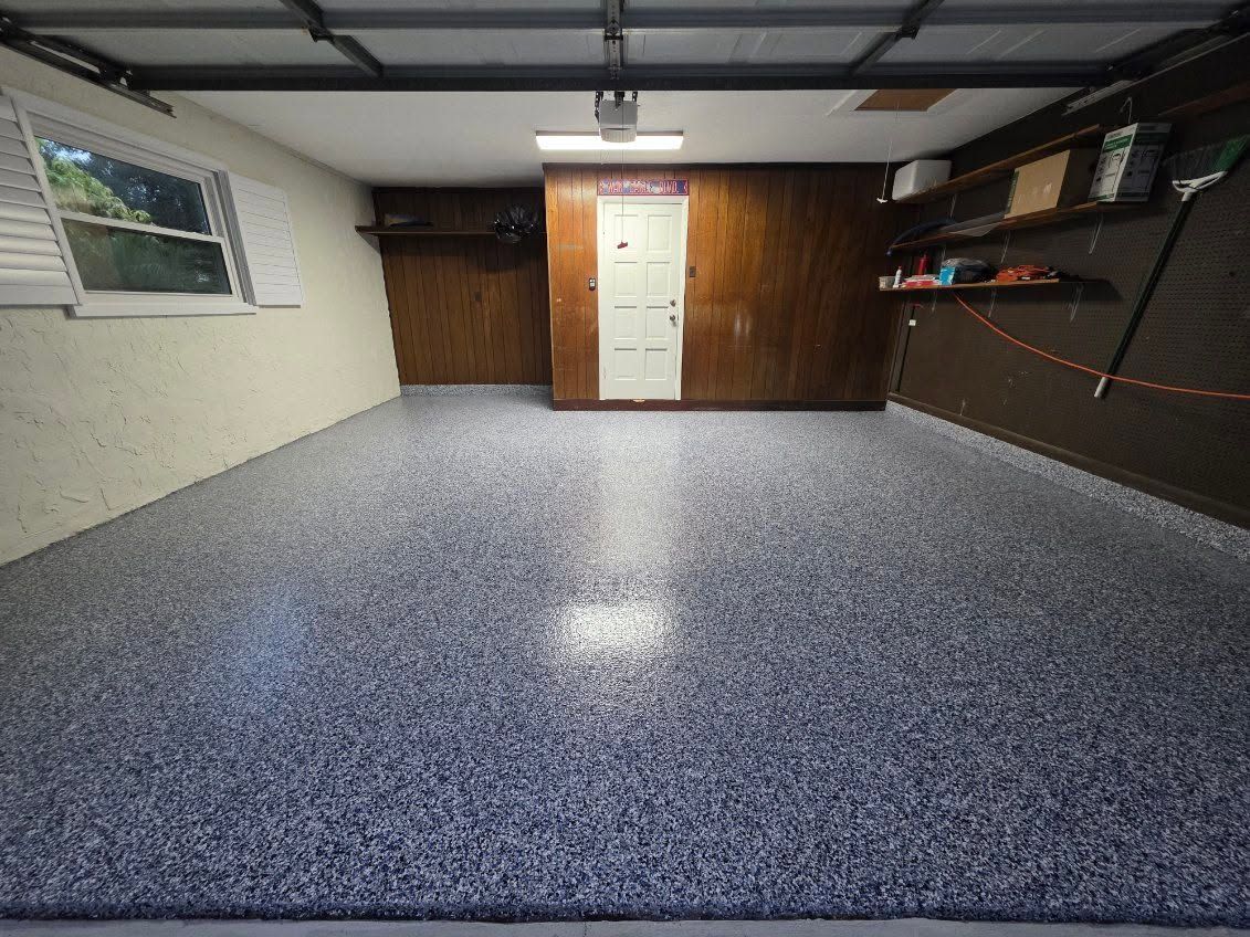 Empty garage with speckled epoxy floor, white door, wood-paneled back wall, and overhead storage racks.