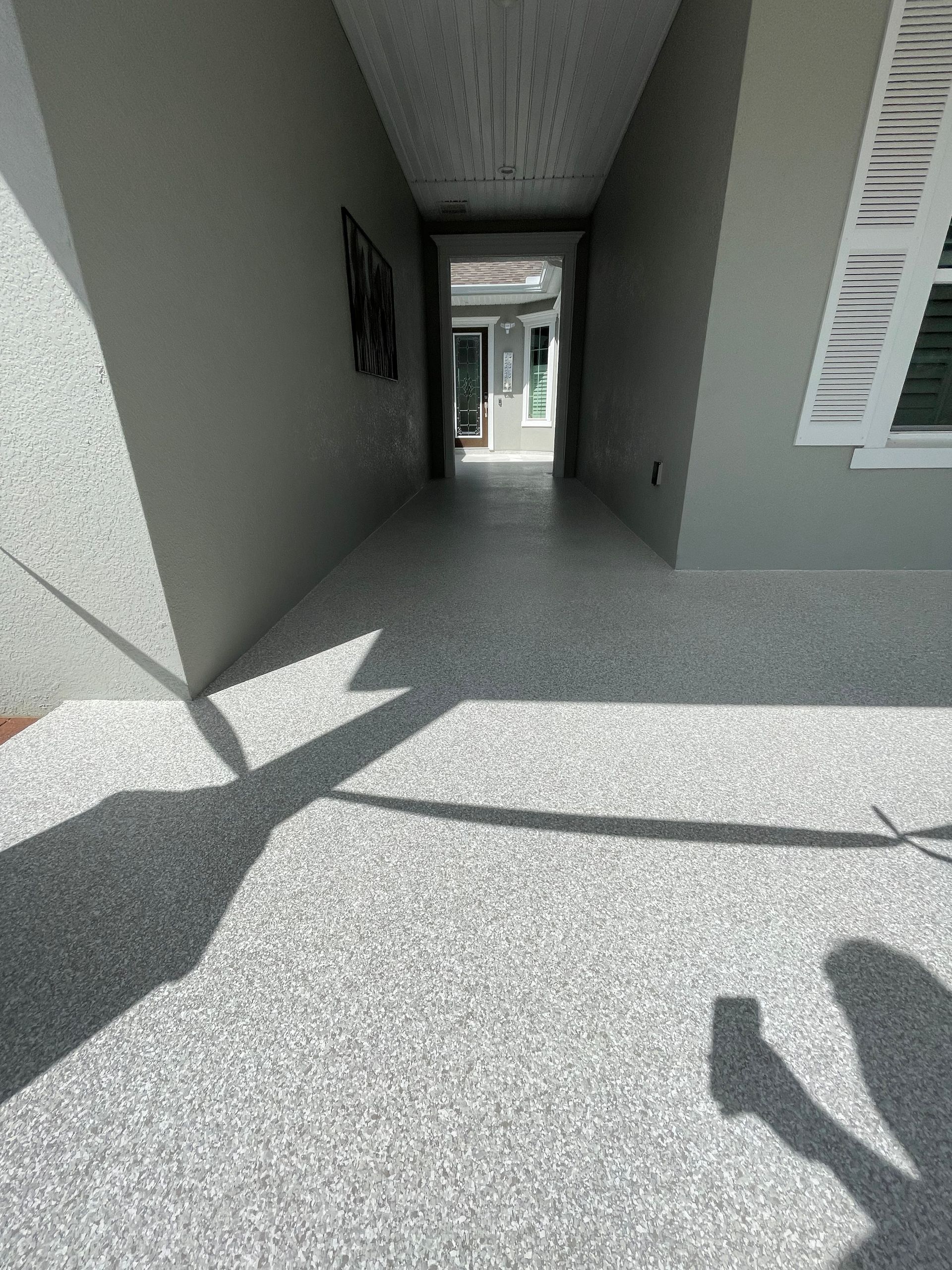 Sunlit covered walkway with gray walls, speckled floor, and a view of the outdoors ahead.