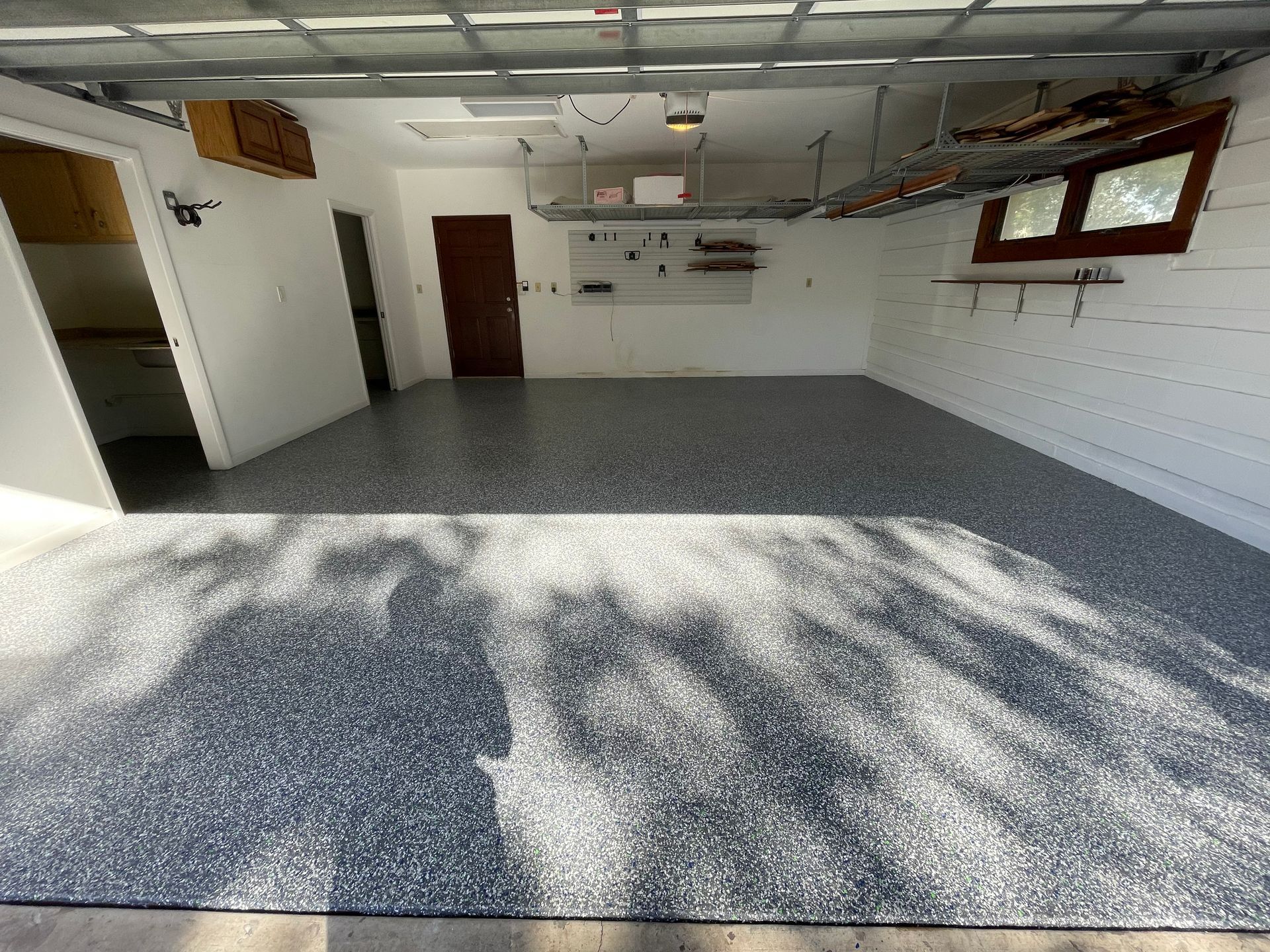 Empty garage with gray speckled floor, white walls, and sunlight casting tree shadows.