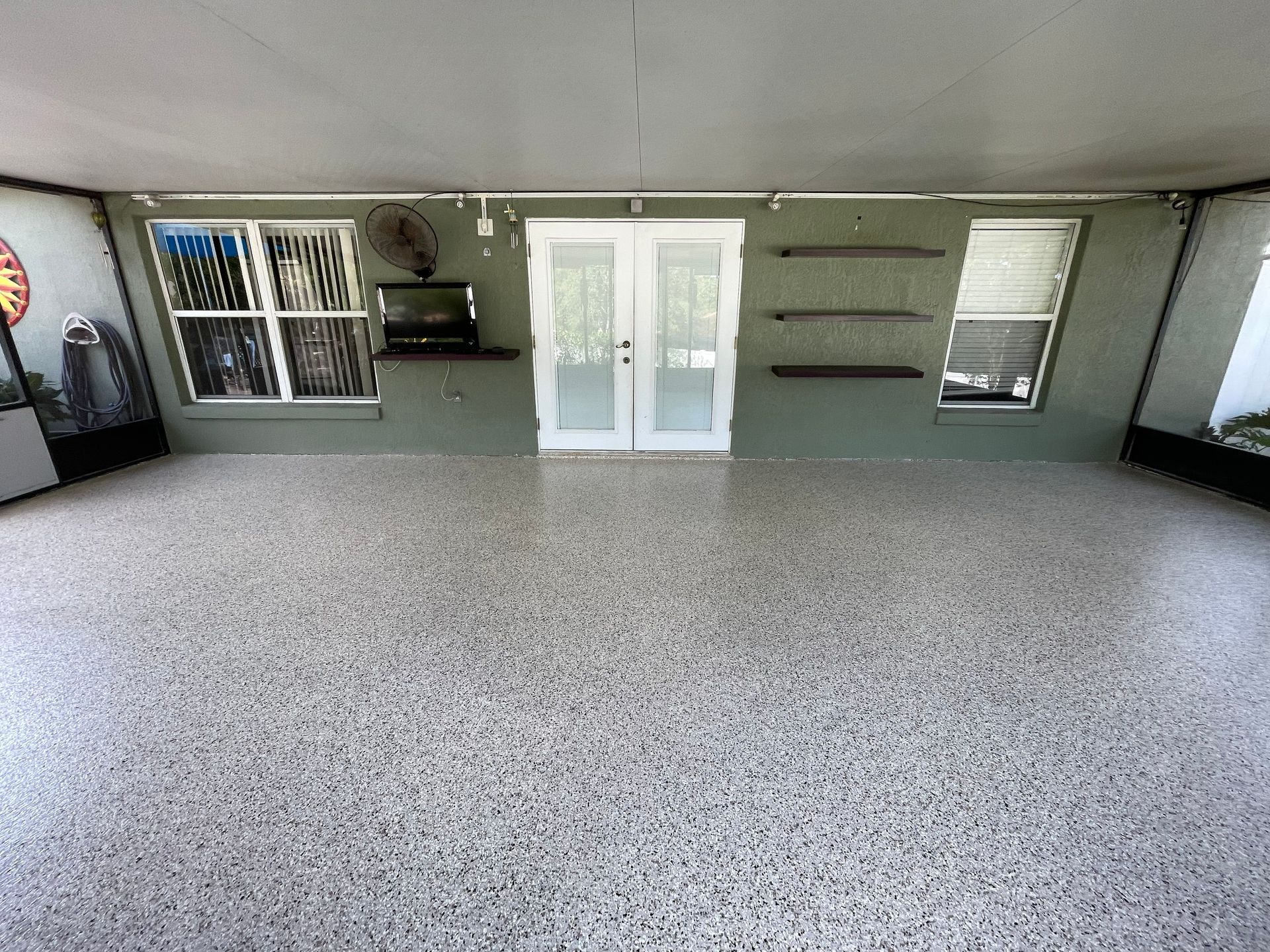 Empty garage with speckled floor, white double doors, shelves, and windows along the back wall