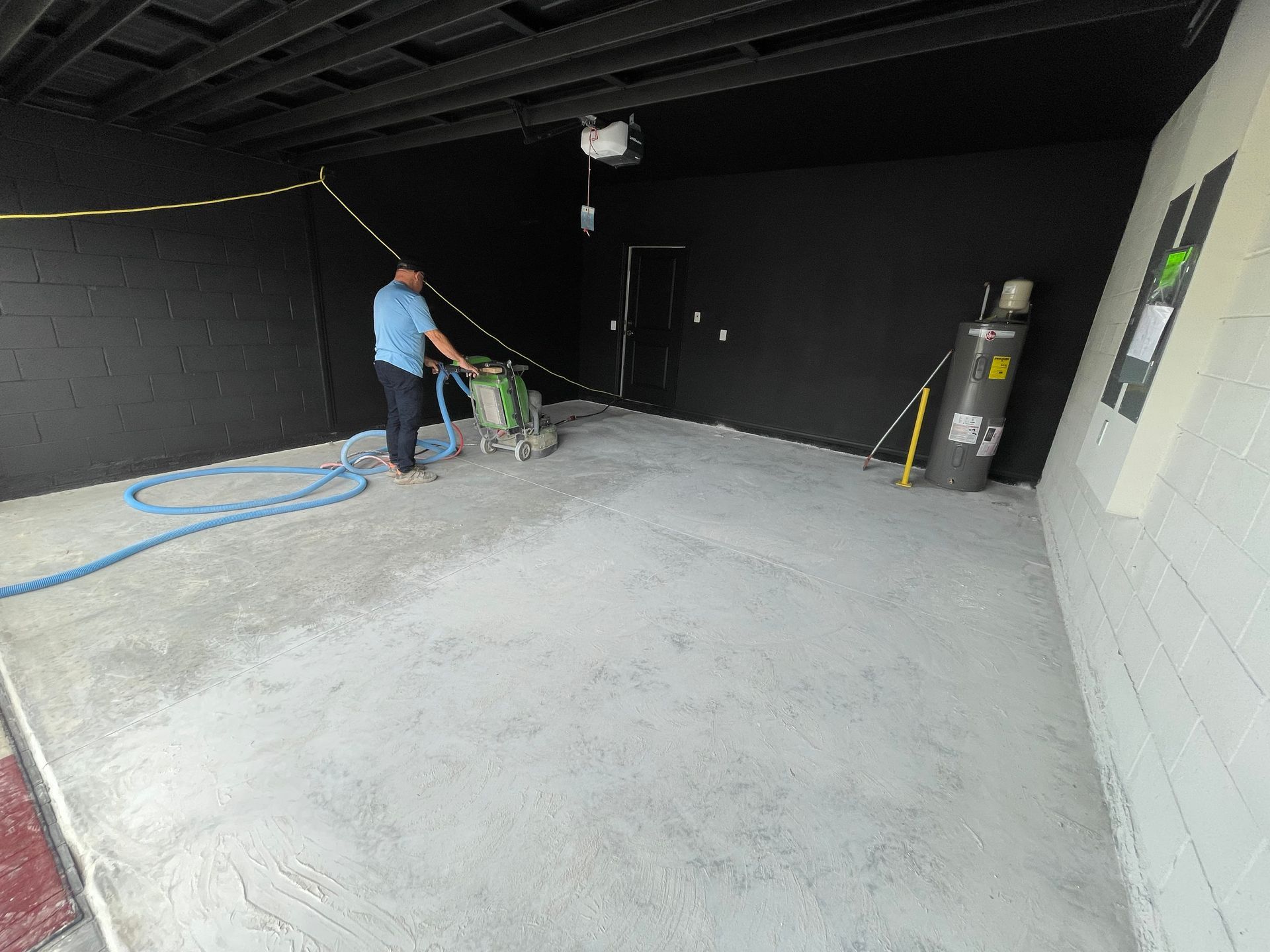 Worker using a floor grinder in an unfinished garage with a concrete floor and dark ceiling