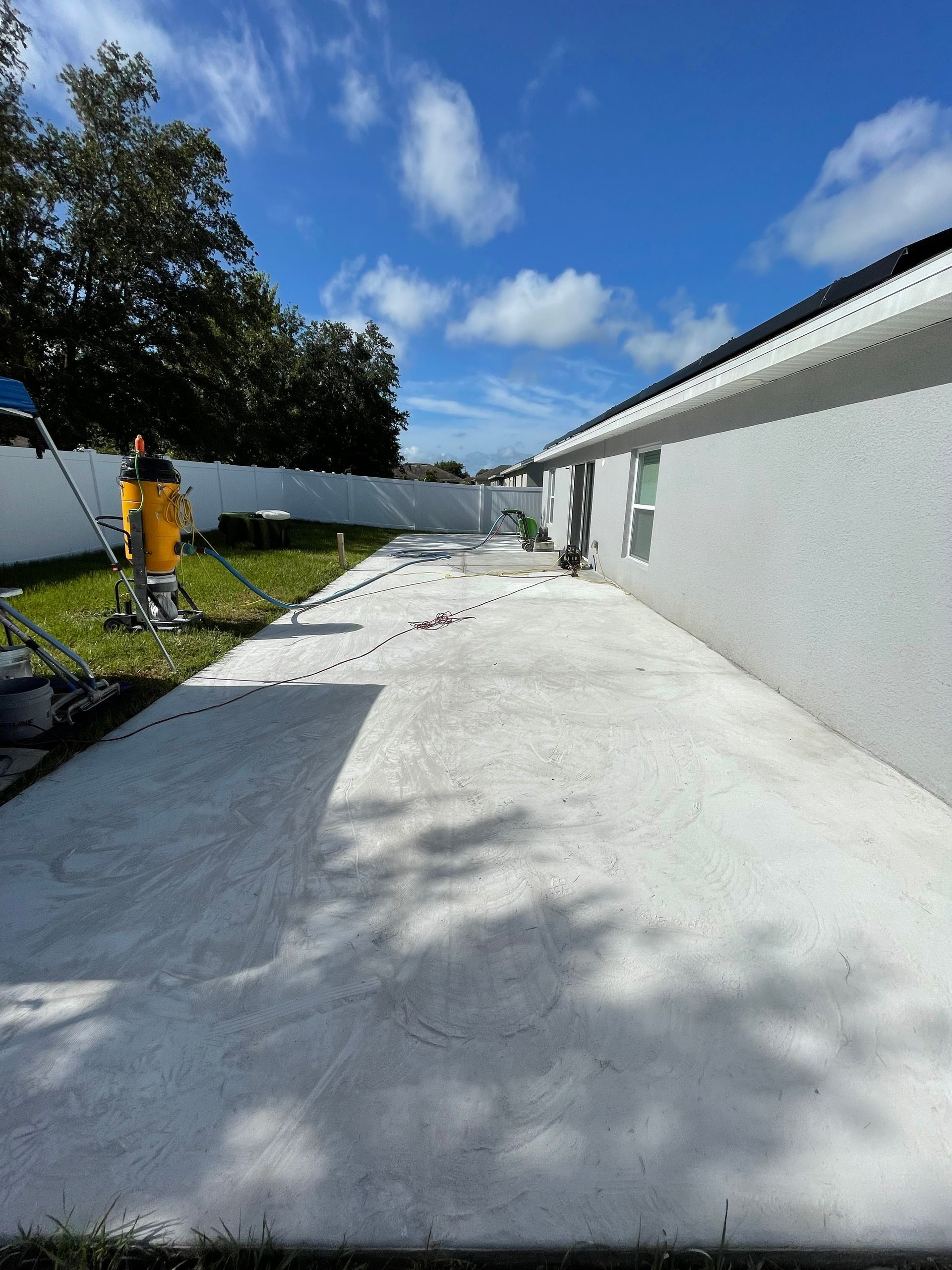 White concrete walkway beside a white building and fenced yard under a blue sky with a yellow machine.