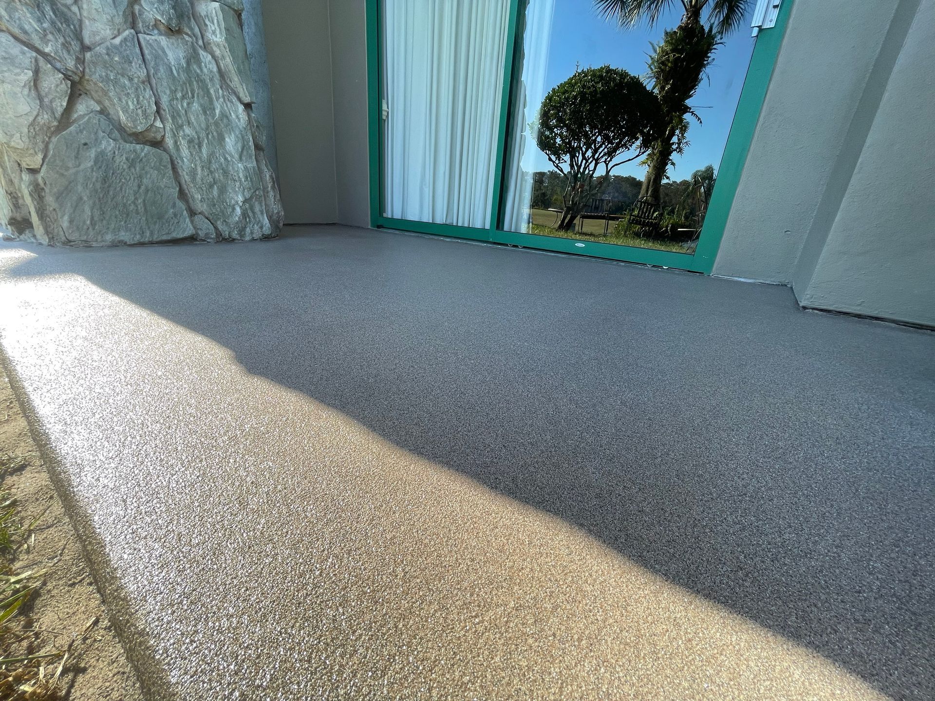 Sunlit textured patio beside a stone wall, with green-framed glass doors opening to a lawn.