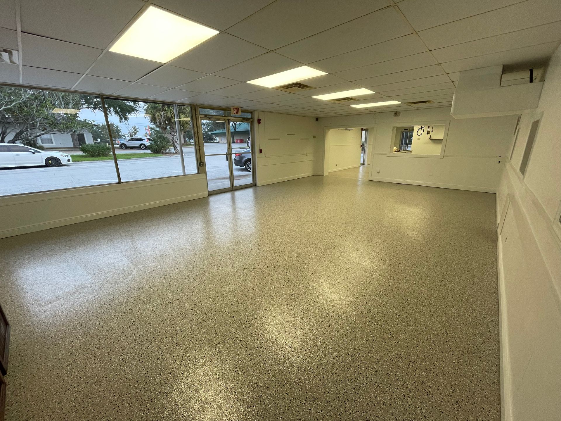 Empty office room with speckled floor, fluorescent lights, and large windows facing outside.