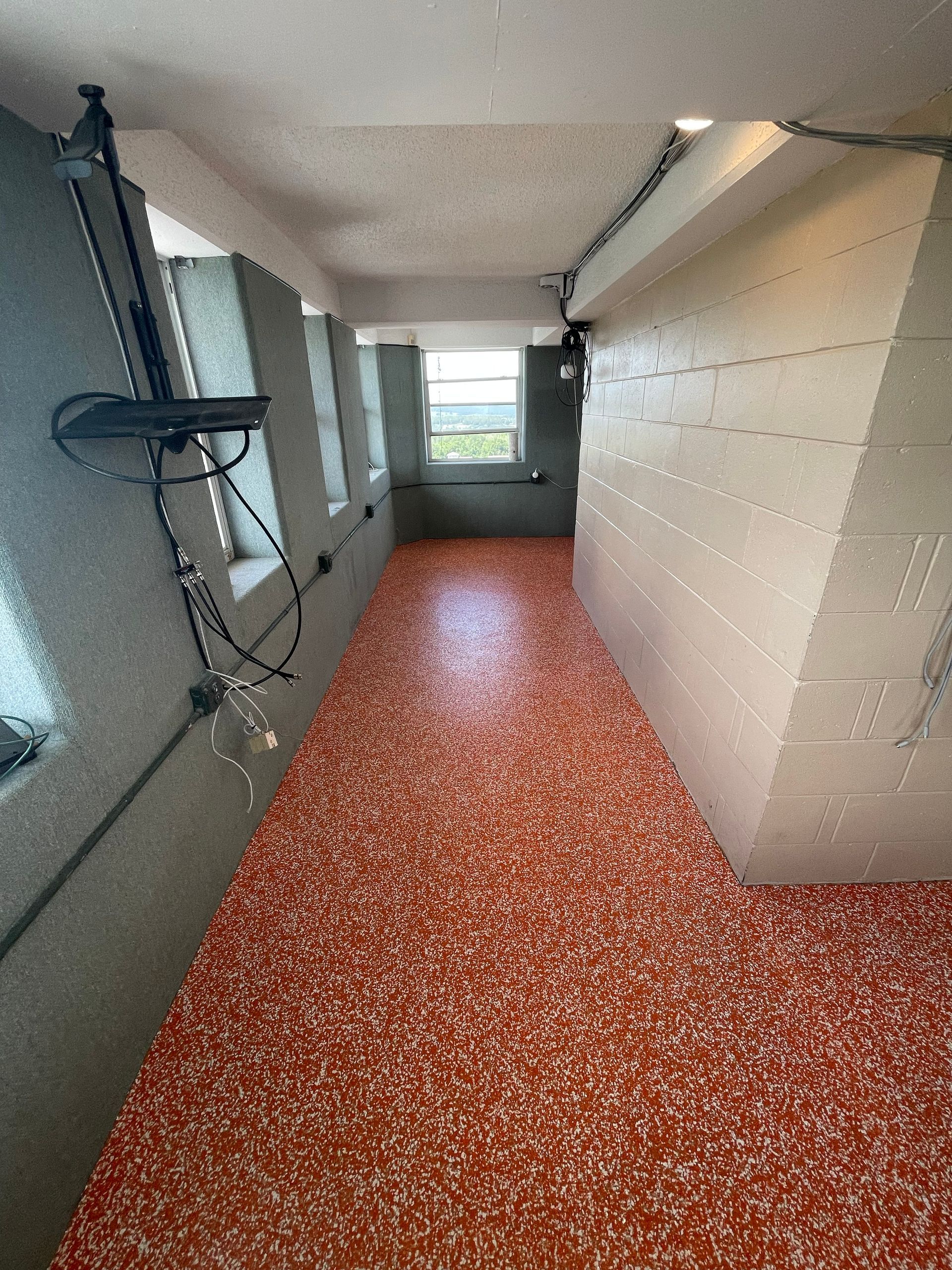 Narrow hallway with red-speckled floor, gray walls, and a window at the end.