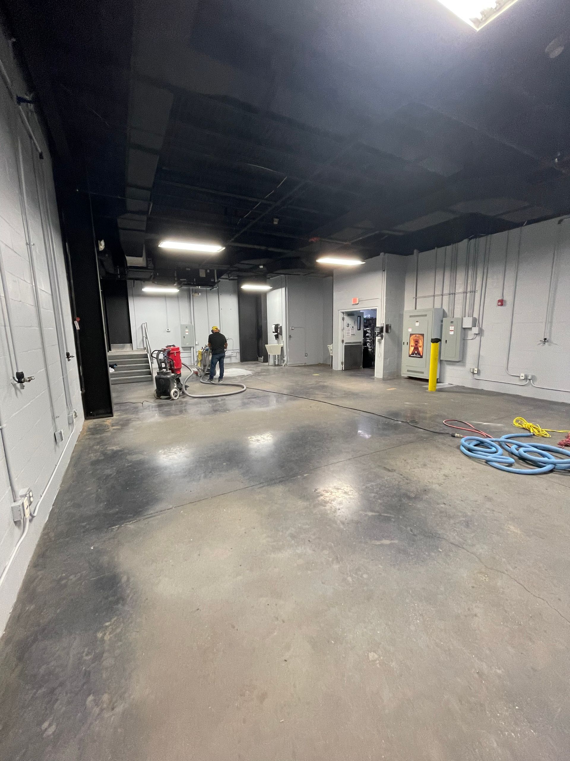 Large empty industrial room with wet concrete floor, overhead lights, and a few people in the distance