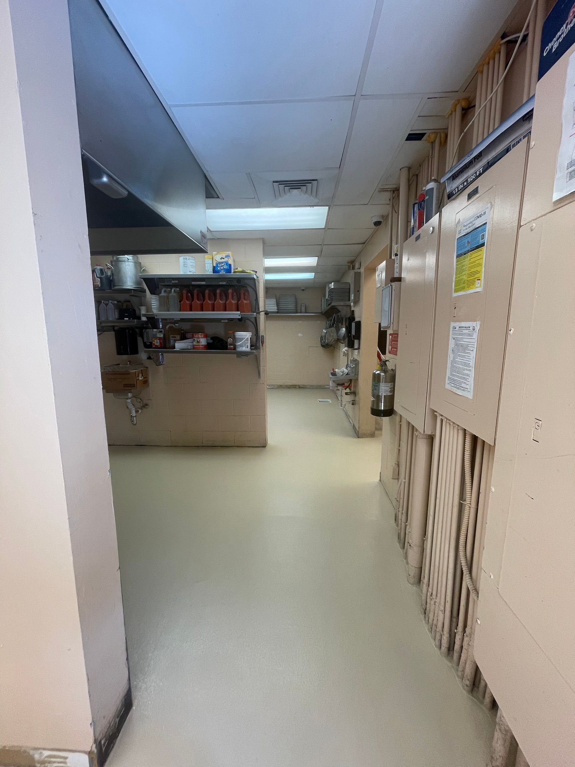 Narrow office hallway with beige walls, fluorescent lights, and a kitchenette at the far end.