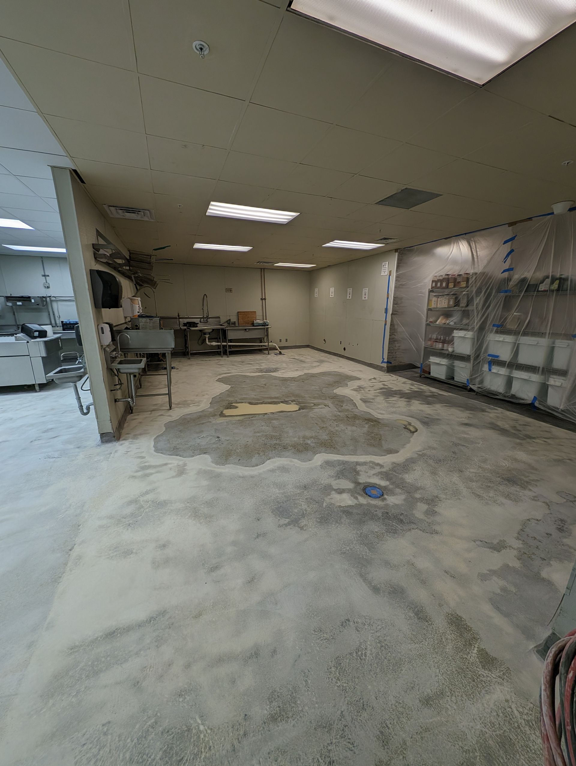 Empty office with damaged, patchy concrete floor and exposed ceiling tiles under fluorescent lights