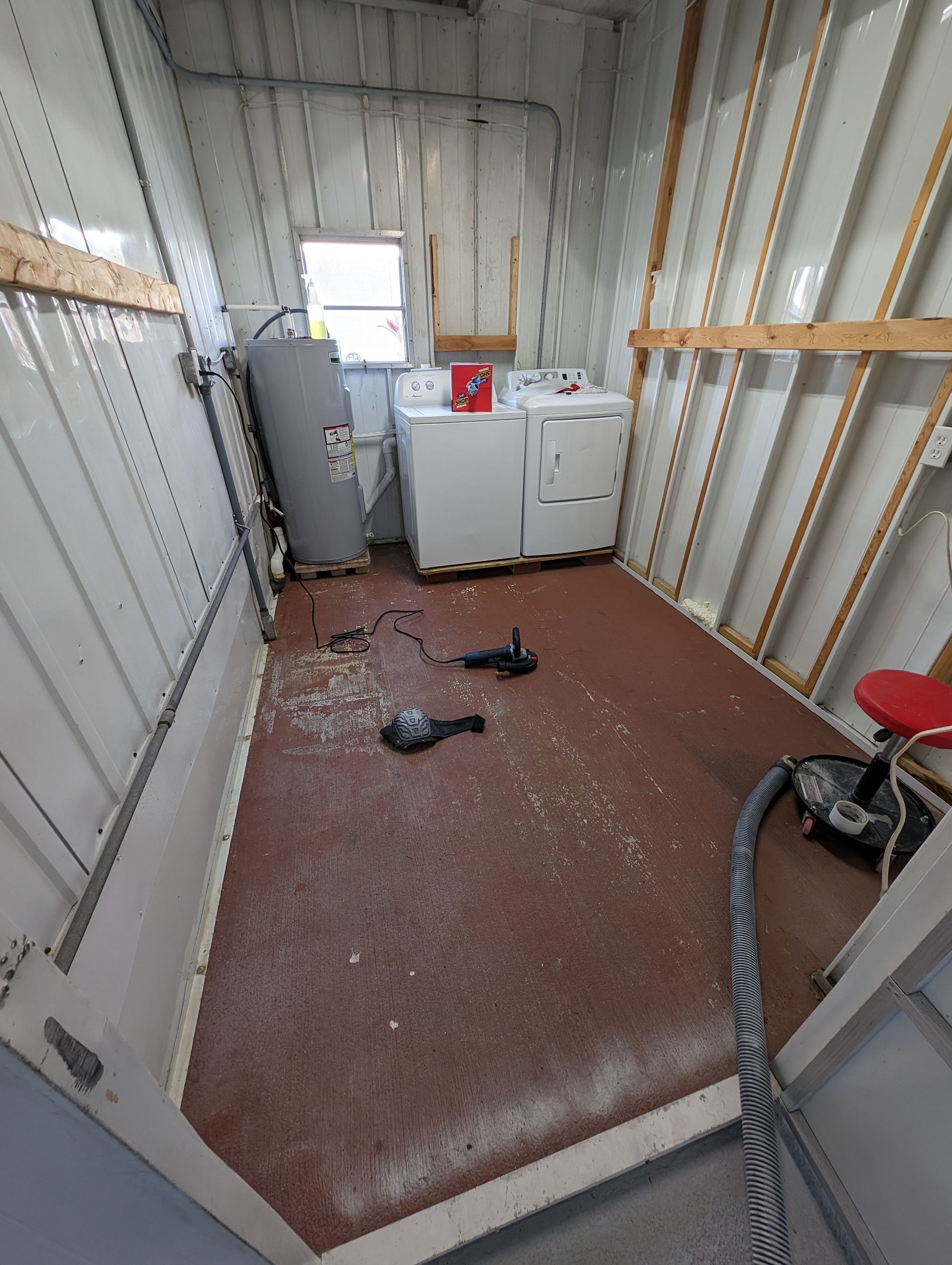 Narrow storage room with white cabinets, a red floor, scattered debris, and a vacuum hose on the right.
