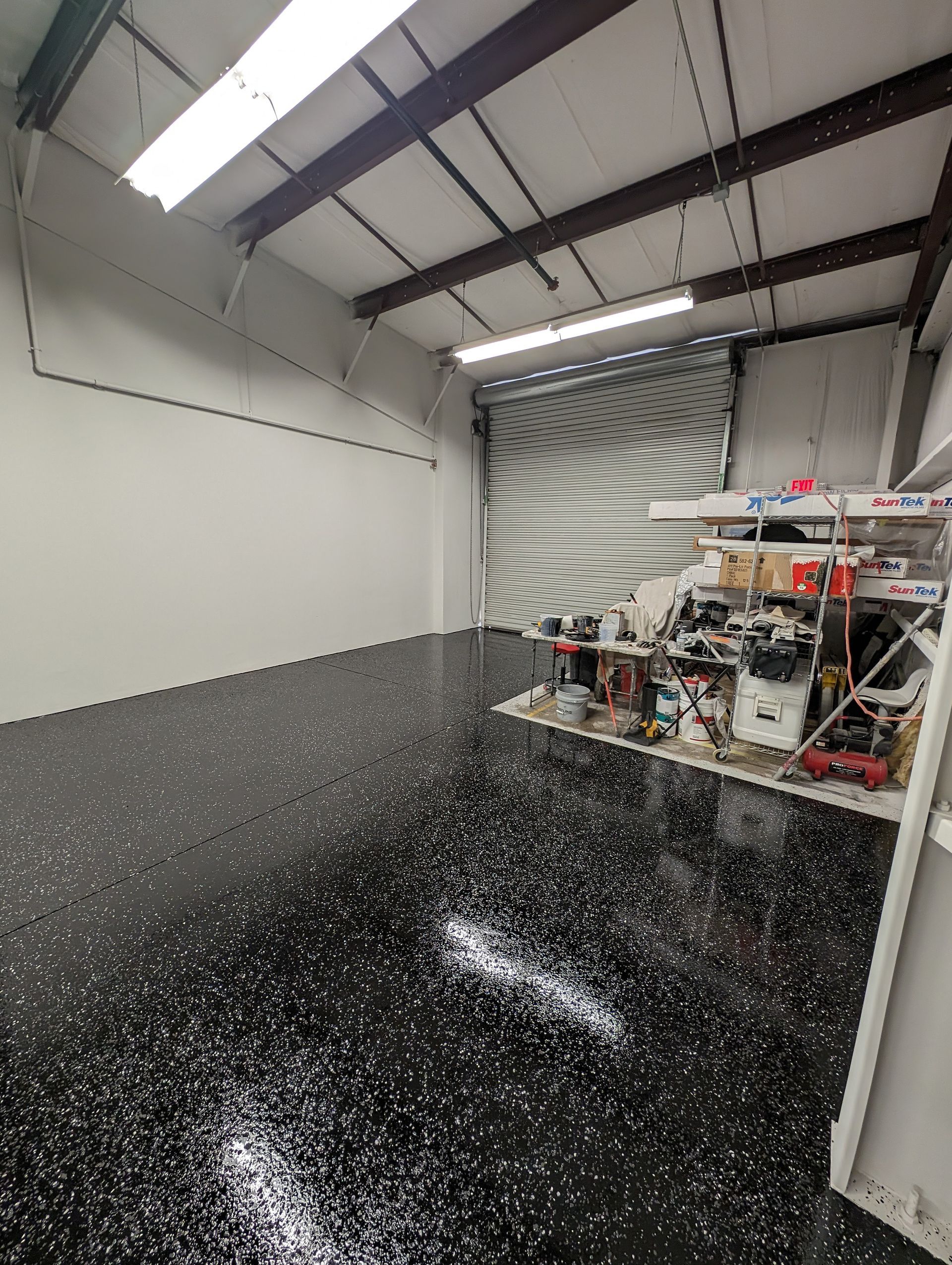 Small workshop with white walls, black speckled floor, overhead lights, and tools near a roll-up garage door