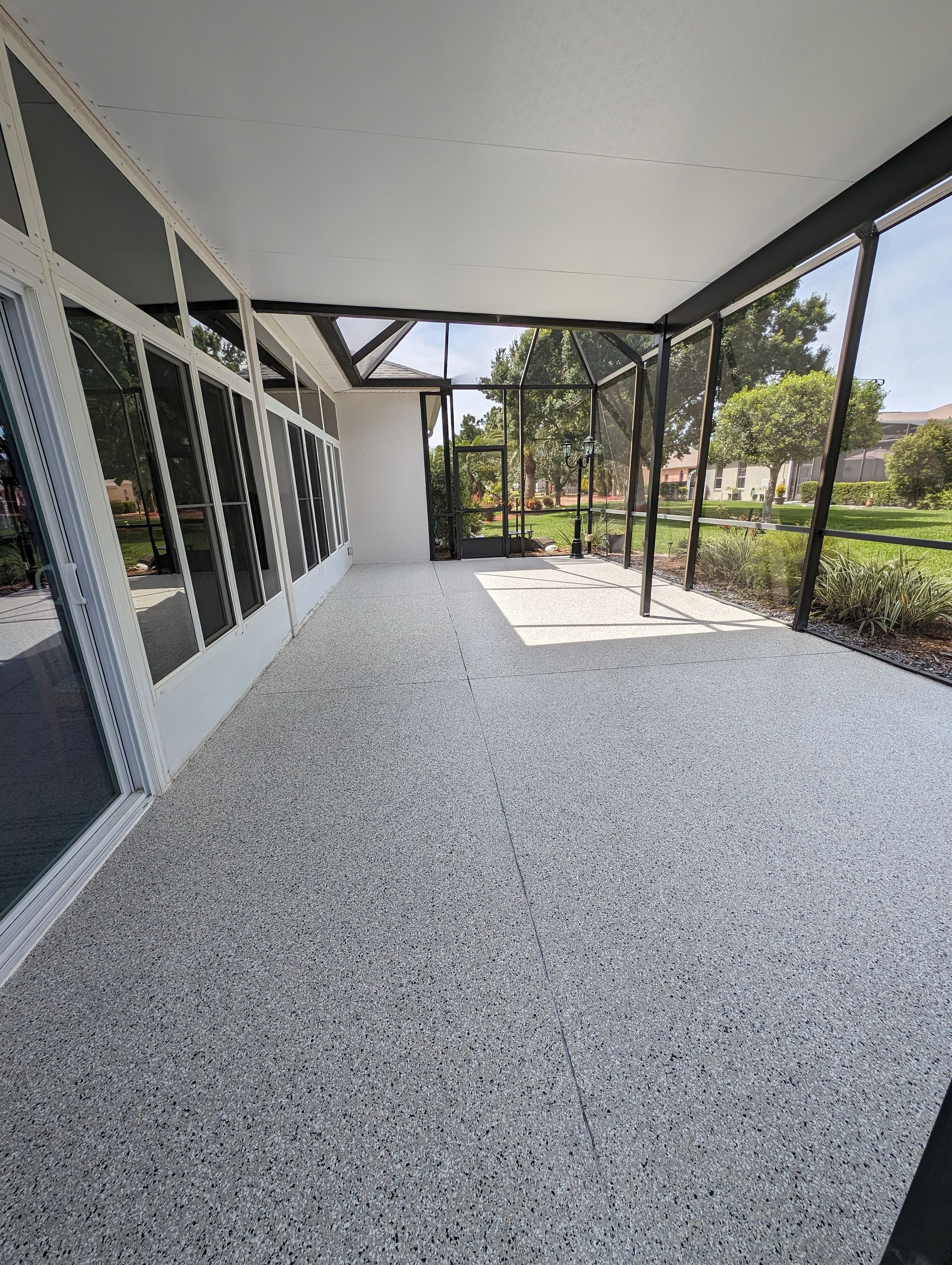 Covered outdoor patio with speckled floor, white wall, and black metal frame overlooking a garden