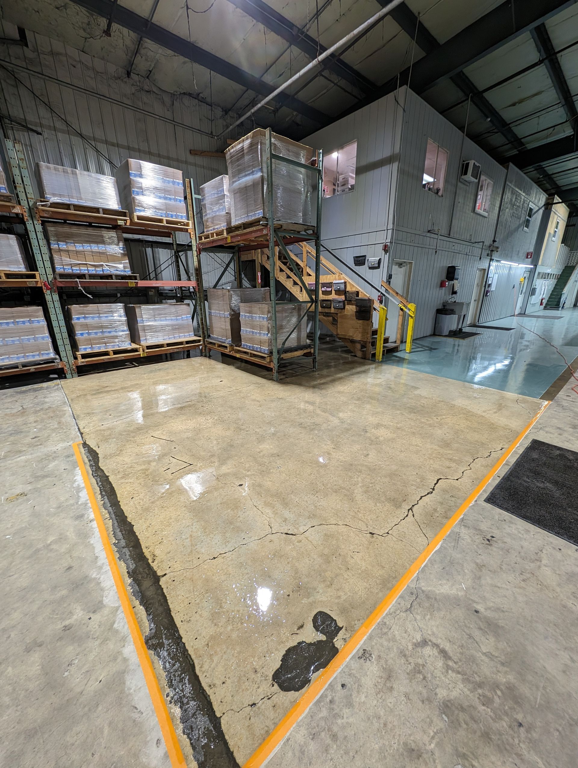 Warehouse interior with stacked pallets, metal stairs, and a polished concrete floor marked by yellow safety lines