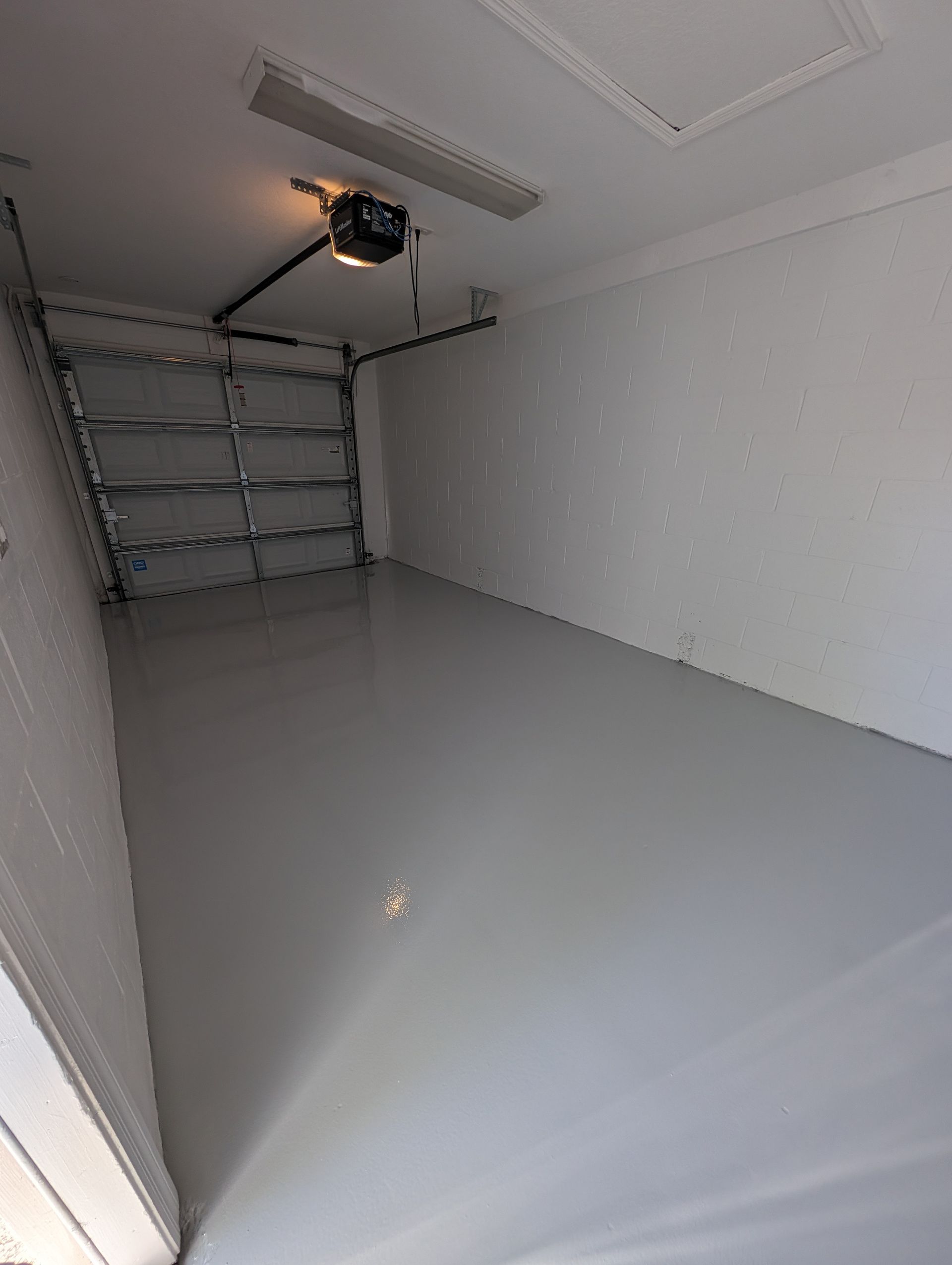 Empty white garage with gray floor, metal shelving on the left, and an open ceiling light fixture.
